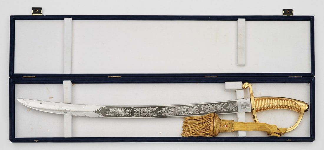 Dekorativer Champagnersäbel: Leicht gebogte, ungeschliffene Klinge mit beidseitig geätztem Blatt- und Bandelwerk, der Darstellung einer Nike mit Lorbeerkranz sowie mehrerer Trophäenbündel. Gerillter Griff mit goldener Zierquas