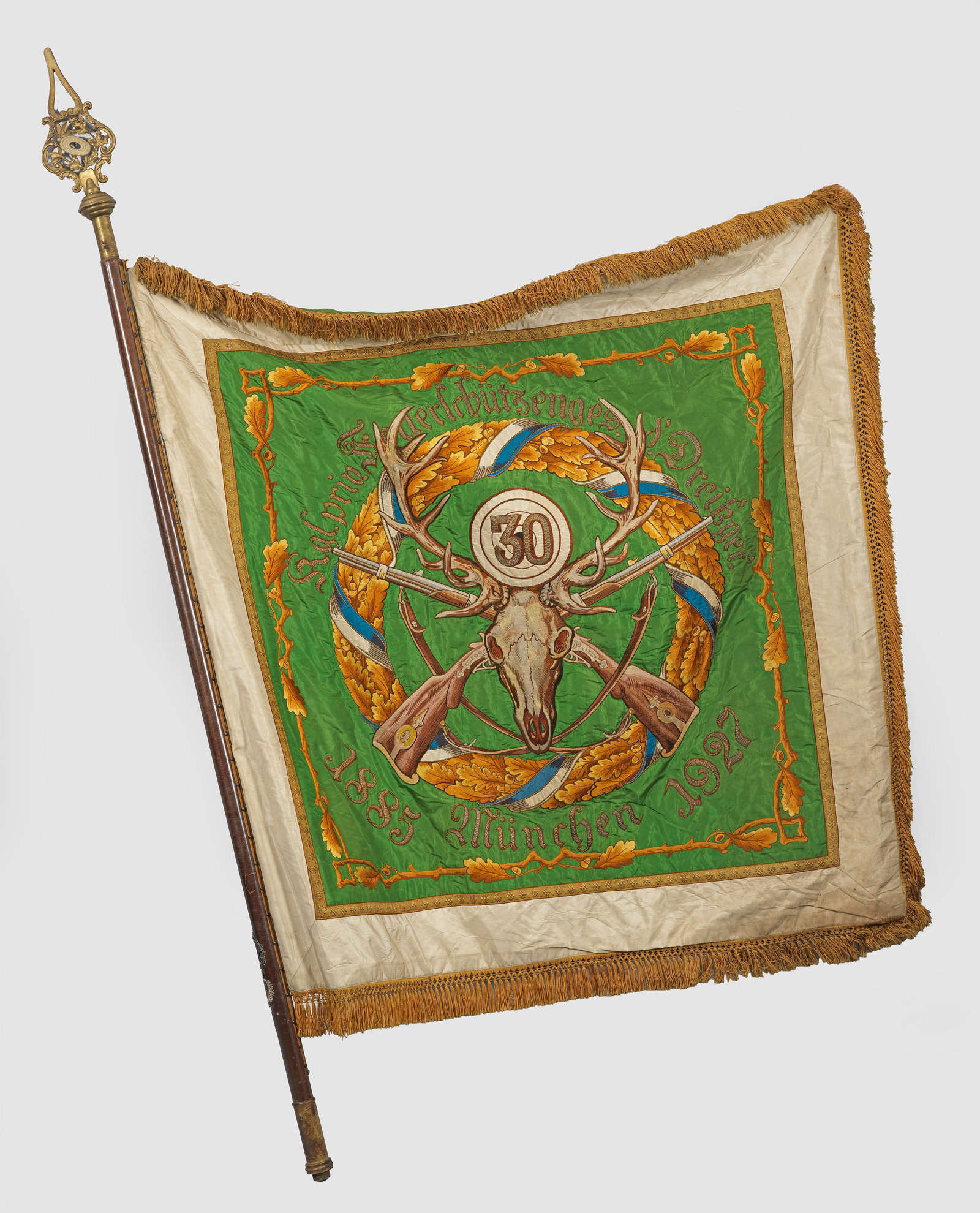 Münchener Schützenfahne von 1927: Holzstange mit durchbrochen gearbeiteter, Jagd-dedizierter Messingspitze. Daran rechteckige, von Fransen gesäumte Form. Beidseitig reiche, polychrom bestickte Jagdmotive. Zentral auf leuchtend grüne