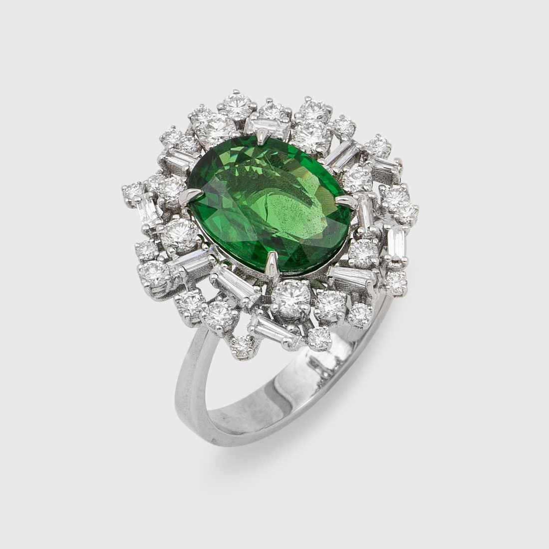 Repräsentativer Entouragering mit Tsavorit: Weißgold, gest. 750. Zentral besetzt mit leuchtend grünem Tsavorit von ca. 3,32 ct. im Ovalschliff, entouriert von Brillanten und Diamantbaguetten von zus. ca. 0,93 ct.; Gew. ca. 5,99 g. Beiliegend 