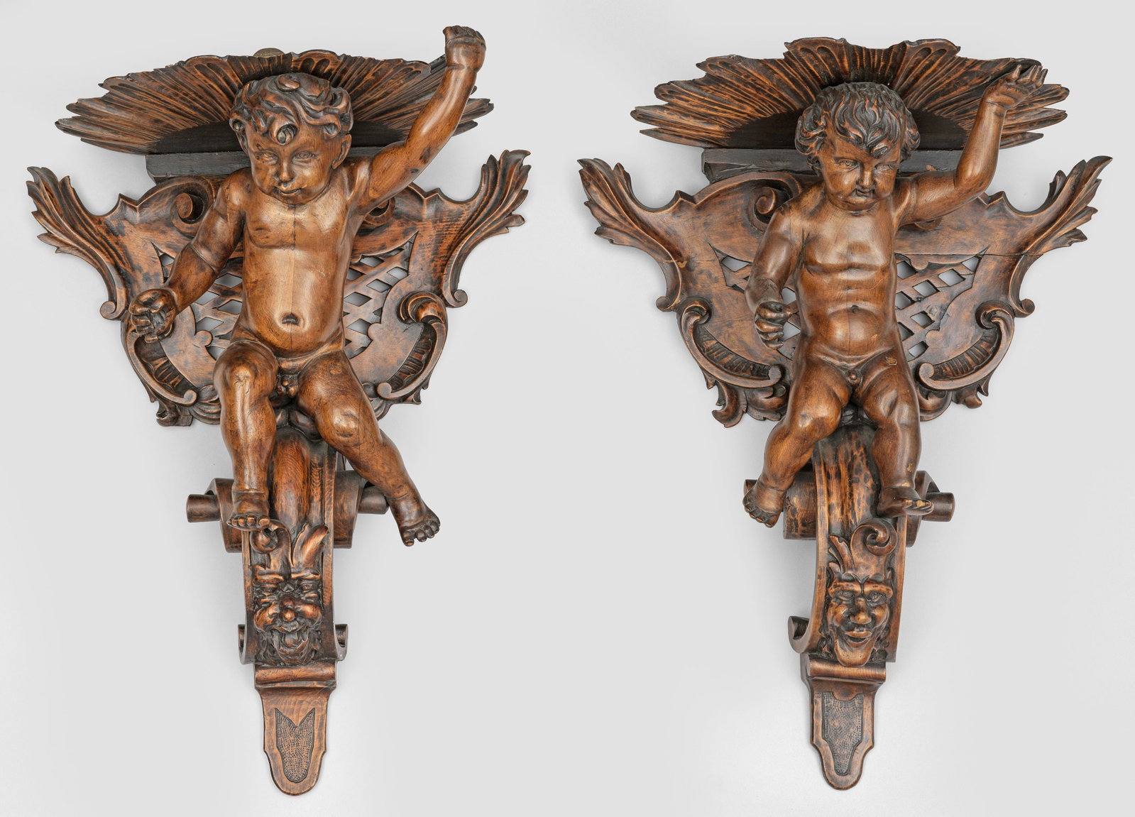 Paar Wandkonsolen im Barock-Stil: Holz, geschnitzt und nussbaumfarben gebeizt. Kartuschenförmiges, gitterartig durchbrochenes Wandschild sowie vollplastische Darstellung eines Puttenkindes auf großer eingerollter Volute mit Fratzenm