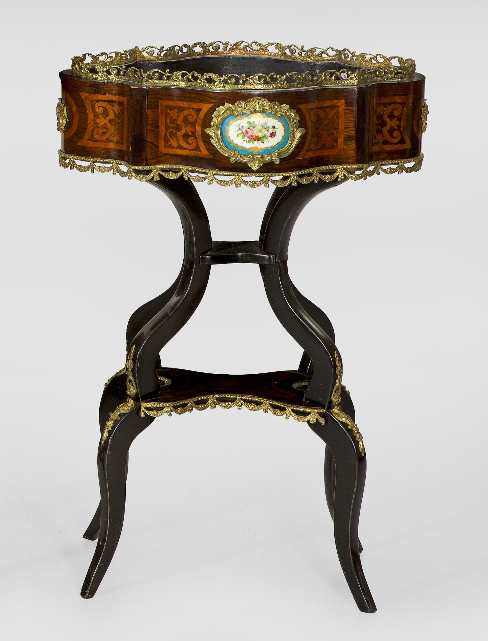 Napoleon III-Jardinière: Palisander und Rosenholz, furniert sowie Applikationen aus Bronze und polychrom gestaltete Porzellanplaketten. Über stark geschwungenen Streben bombierter, kartuschenförmiger Korpus mit durchbrochen