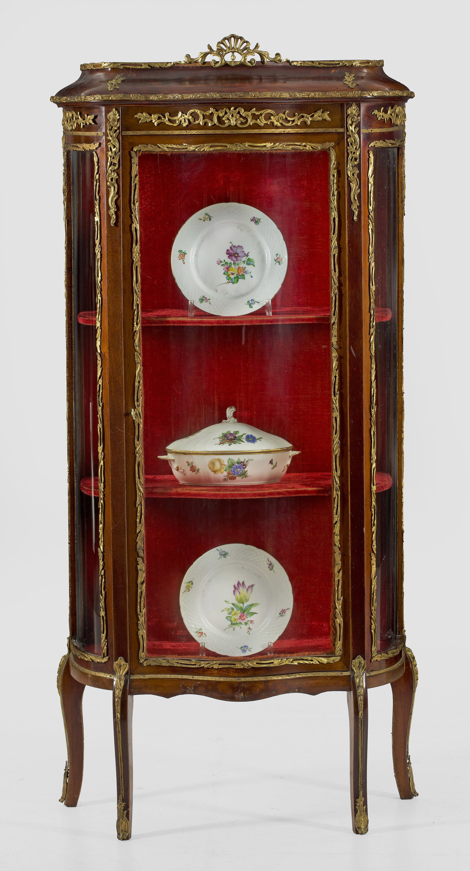 Napoleon III-Salonvitrine: Mahagoni, furniert. Dreiseitig bombierter, verglaster Korpus auf geschweiften Beinen. In der Front hohe Vitrinentür. Geschweift ansteigender oberer Abschluss mit durchbrochen gearbeiteter Muschelbekr