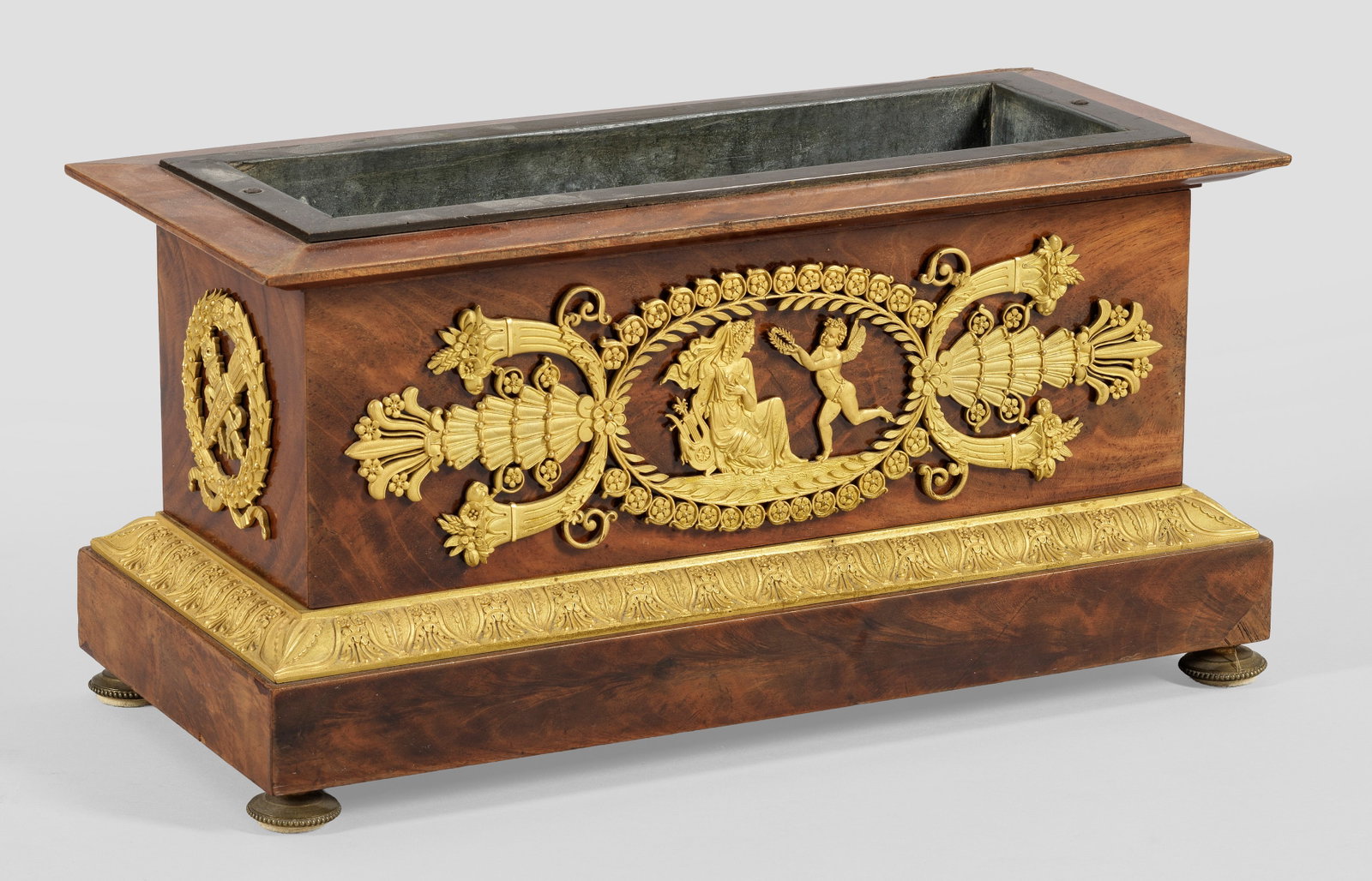Kleine Jardinière im Empire-Stil: Mahagoni, furniert sowie Applikationen aus vergoldeter Bronze. Gerader, querrechteckig angelegter Korpus auf schmalem Sockel mit Palmettenfries und gedrückten Füßen. Allseitig reiche, teils figural