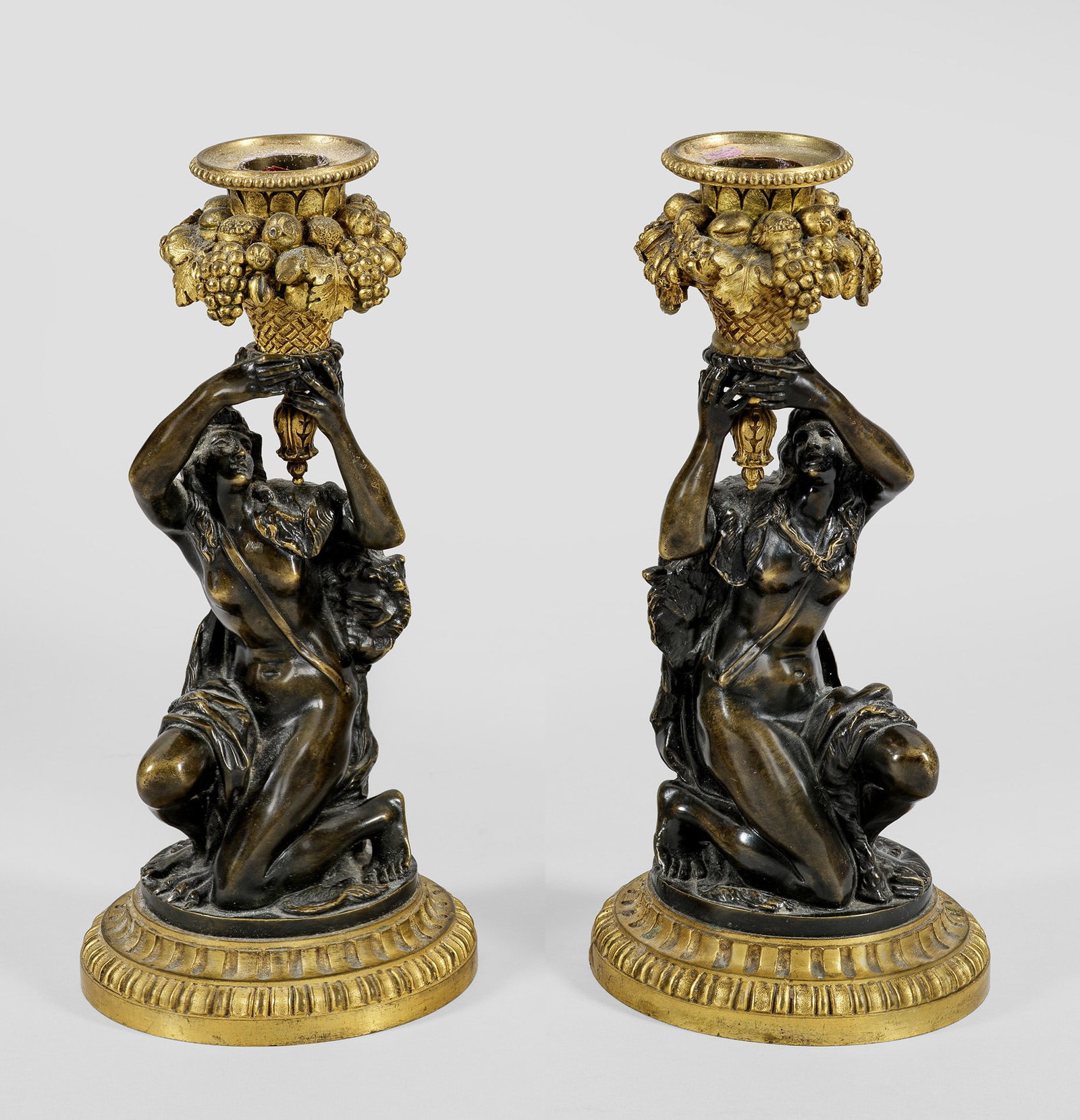 Paar Napoleon III-Kerzenleuchter: Bronze, patiniert und teilw. vergoldet. Auf getrepptem Rundsockel kniende, mit einem Löwenfell bekleidete Frauenfigur, eine Tülle in Form eines mit Früchten gefüllten Füllhorns haltend. H. 22 cm.