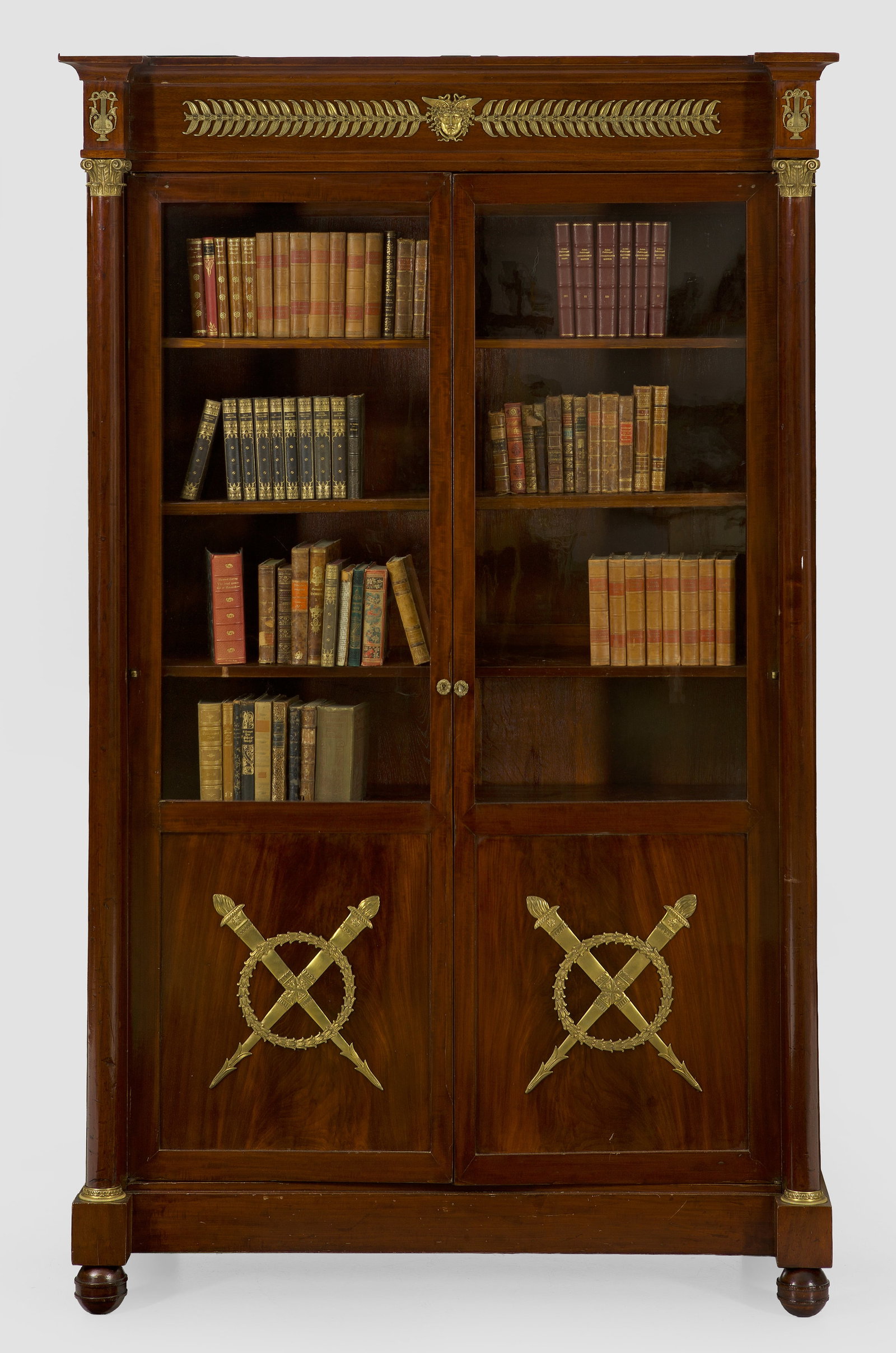 Empire-Bücherschrank: Mahagoni, furniert sowie Applikationen aus Bronze. Gerader, hochrechteckig angelegter Korpus auf gerader Zarge und Kugelfüßen. In der gegliederten Front zwei verglaste Türen, flankiert von eingeste
