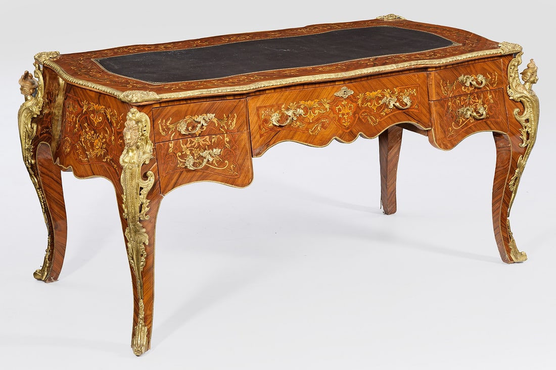 Repräsentatives Bureau plat im Napoleon III-Stil: Nussbaum furniert sowie farbige Einlagen. Allseitig bombierter Korpus, flankiert von weiblichen Genien auf geschwungenen Beinen. In der gegliederten Front drei Schubladen unterschiedlicher Größe.