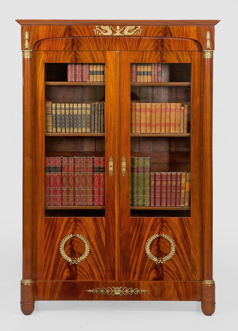 Bücherschrank im Empire-Stil: Mahagoni, furniert. Gerader, hochrechteckig angelegter Korpus auf geradem Zargensockel und gerundeten Füßen. In der gegliederten Front zwei verglaste Vitrinentüren, flankiert von eingestellten Säu