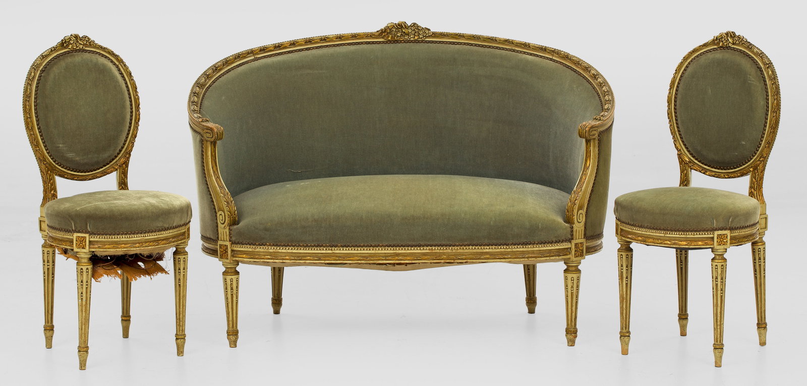 Salongarnitur im Louis XVI-Stil: 3-tlg.; Canapé und zwei Stühle. Holz, geschnitzt und cremegrün gefasst. Bombierte Zarge auf schlanken, kannelierten Balusterbeinen. Blattreliefierte Stützen für geschweift ansteigende, bombierte 