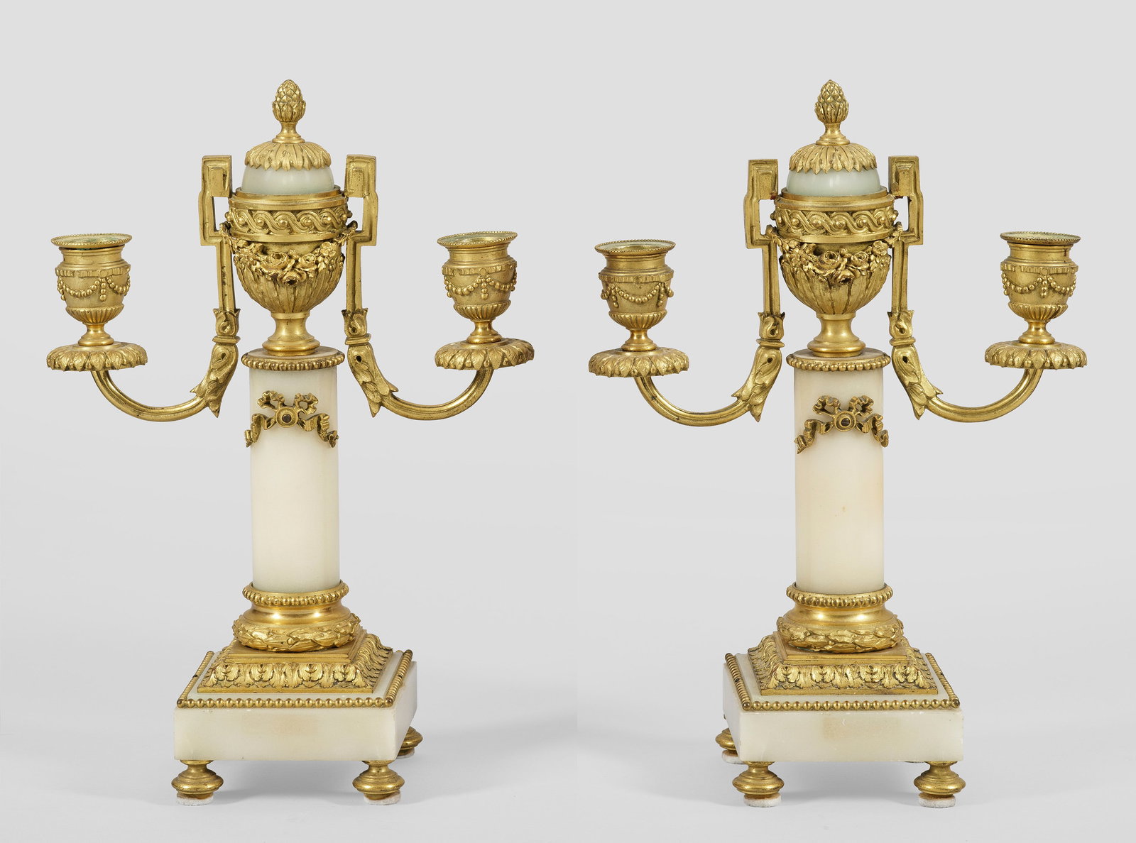 Paar Girandolen im Louis XVI-Stil: 2- bzw. 3-flg.; Cremefarbener Alabaster und vergoldete Bronze. Über quadratischer Plinthe mit Palmetten- und Perlstabfries zylinderförmiger Schaft mit Vasenbekrönung als Pinienknauf. Davon ausgehen
