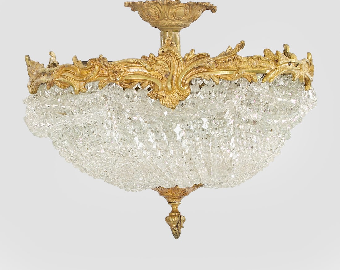 Plafoniere im Louis XV-Stil: 1-flg.; Bronze und farbloses Glas. Kuppelartig angelegter Korpus aus Prismenschnüren im Verlauf. Umlaufend breiter Fries aus reliefplastisch gestalteten Voluten. Elektrifiziert. H. 32 cm. D. 38 cm.