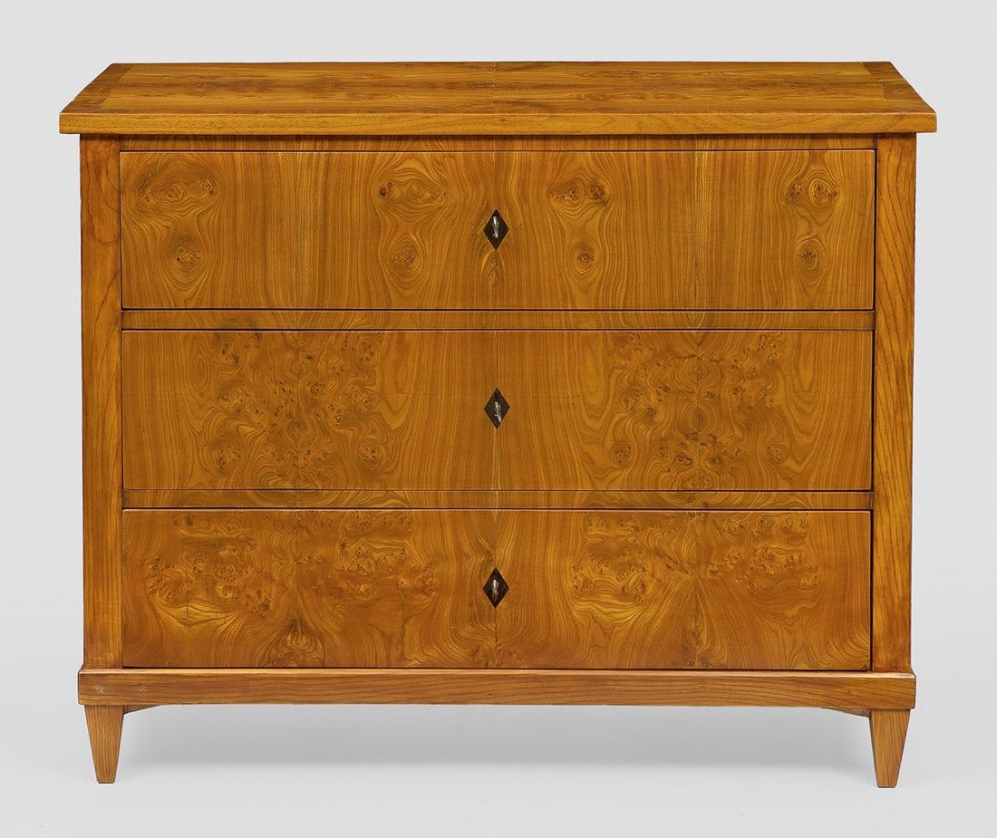 Biedermeier-Kommode: Rüster, furniert. Gerader, querrechteckiger Korpus auf schmaler Zarge und konischen Vierkantbeinen. In der gegliederten Front drei Schubladen. Gering überstehende Deckplatte. H. 82 cm. 103 cm x 50 c