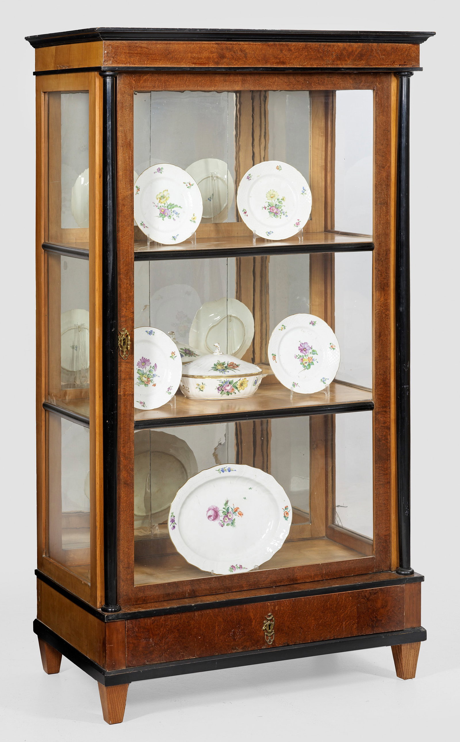 Biedermeier-Vtitrinenschrank: Kirschbaum und Thujamaser, furniert sowie teilw. ebonisiert. Gerader, hochrechteckig angelegter, dreiseitig verglaster Korpus mit einschübigem Zargensockel auf konischen Vierkantfüßen, flankiert vo