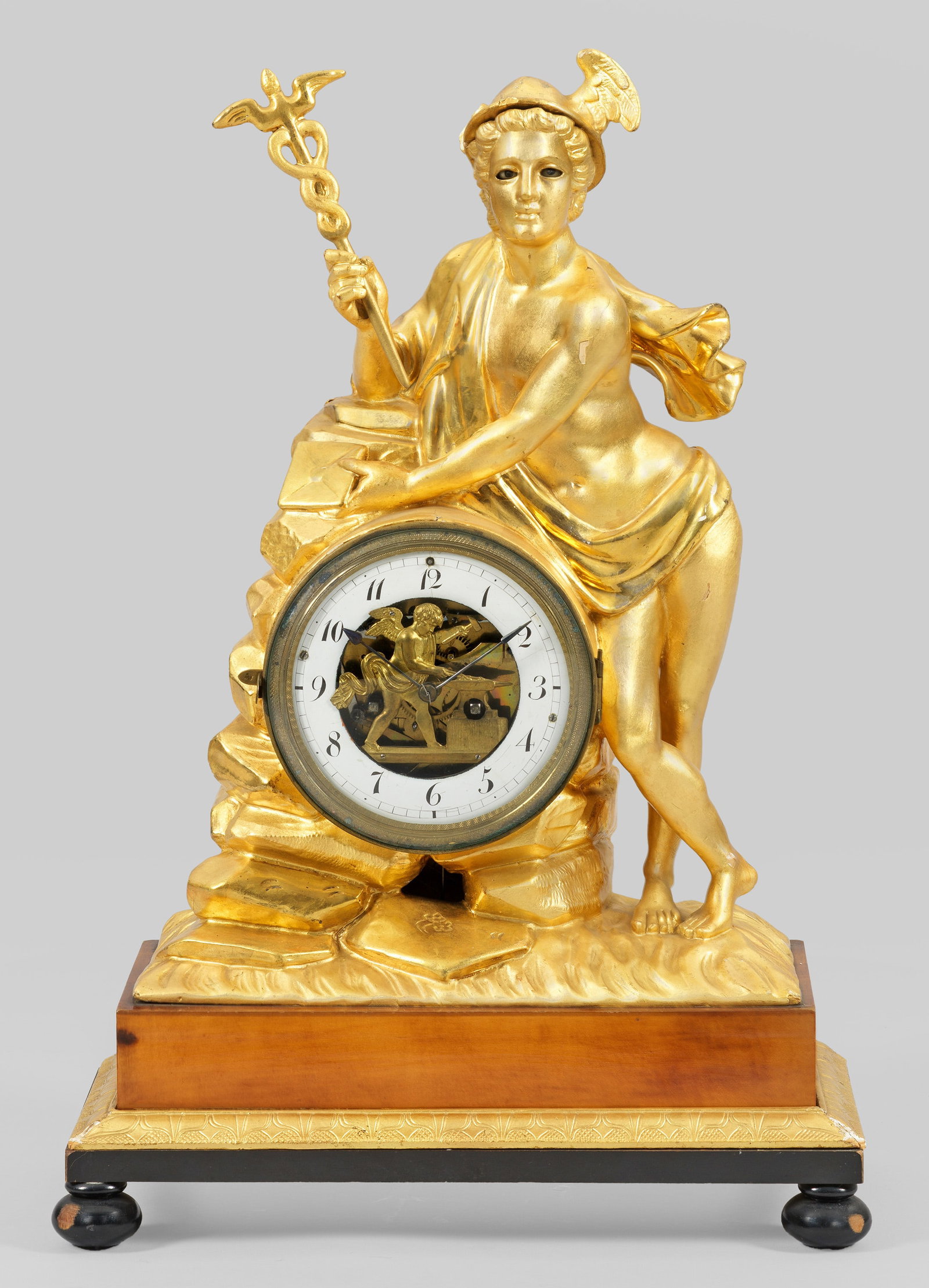 Seltene Wiener Biedermeier-Uhr mit Automatenfunktion: sog. Augenwender-Uhr. Holz, geschnitzt, gefasst und vergoldet sowie Obstholz, furniert. Über querrechteckig angelegter Basis mit stilisiertem Palmettenfries, vollplastische Darstellung Merkurs. Profi
