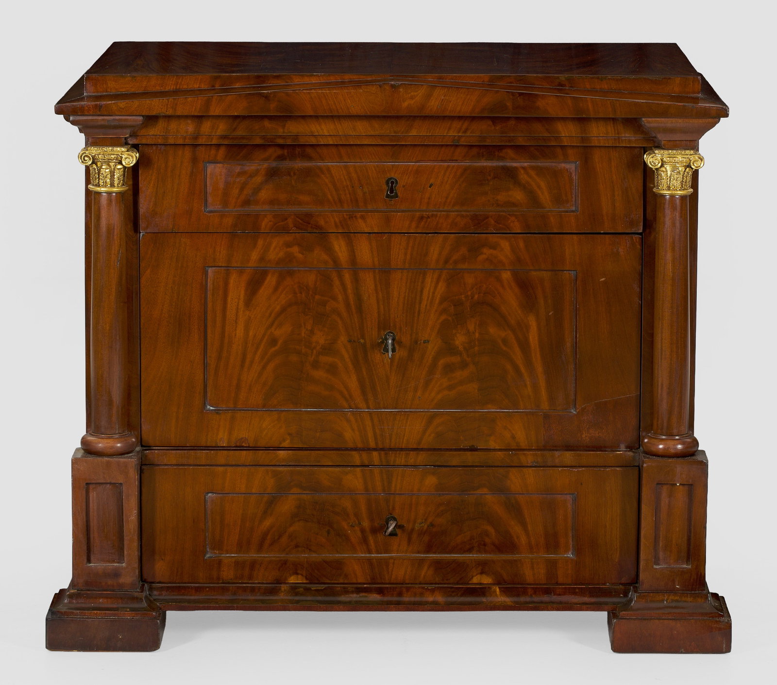 Biedermeier-Kommode: Mahagoni, furniert. Gerader, rechteckig angelegter Korpus, flankiert von eingestellten Säulen auf gekanteten Füßen. In der gegliederten Front drei kassettierte Schubladen unterschiedlicher Größe.