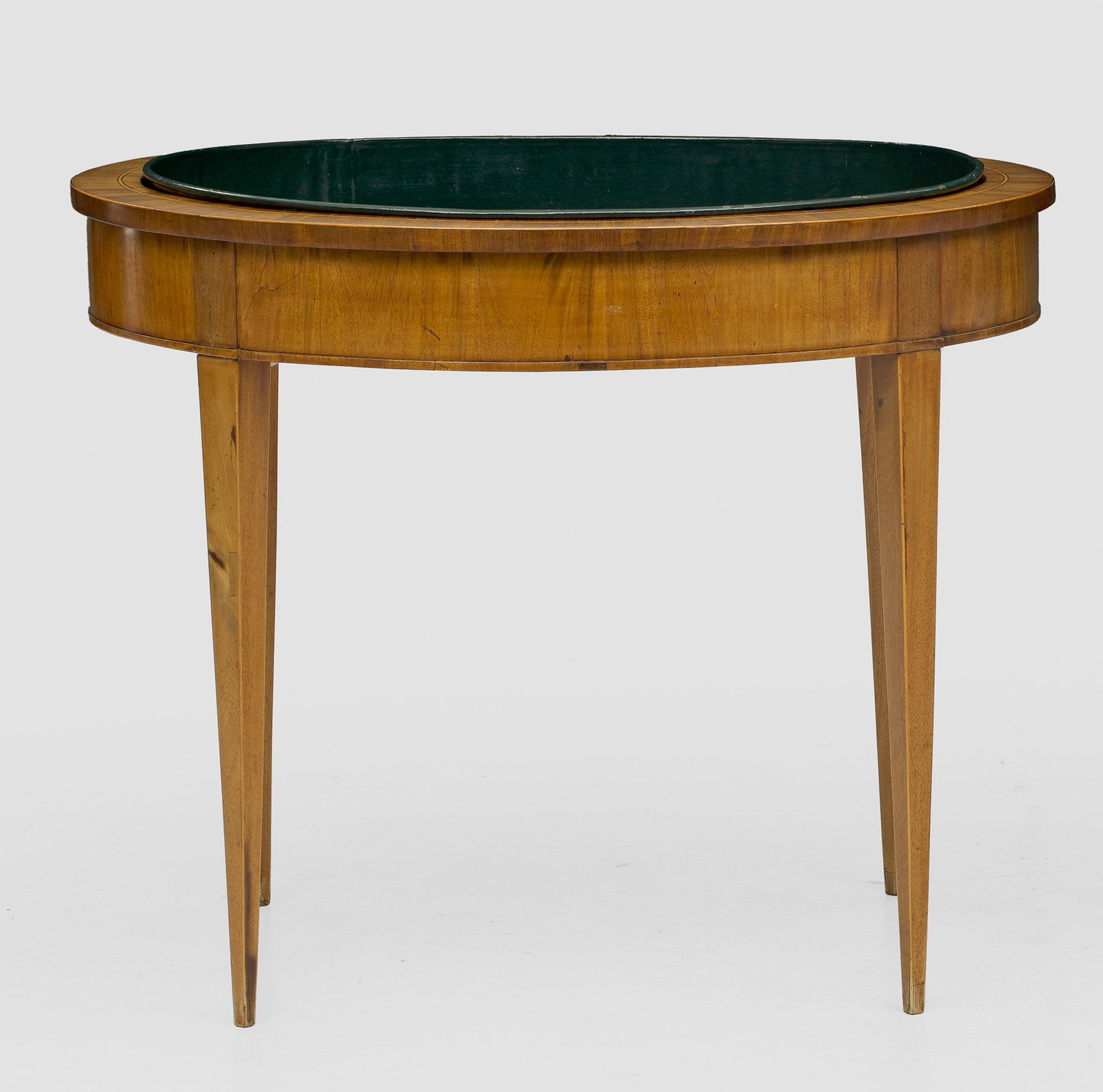 Biedermeier-Jardinièrentisch: Mahagoni, furniert. Bombierter Zargenkasten auf hohen, konischen Vierkantbeinen. Gering überstehende, ovale Deckplatte mit eingesetzter Pflanzschale aus lackiertem Metall. H. 73 cm. 88 cm x 71 cm. A 