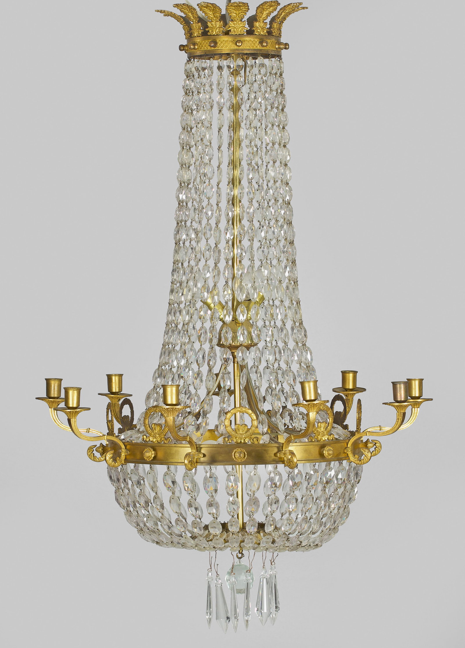 Deckenlüster im Empire-Stil: 9-flg.; Messing und farbloses Glas. Balusterförmiger Korpus aus Prismenschnüren im Verlauf. Schmaler profilierter Reif mit Bekrönungen aus Lorbeerkränzen als Träger für volutenförmige Leuchtera