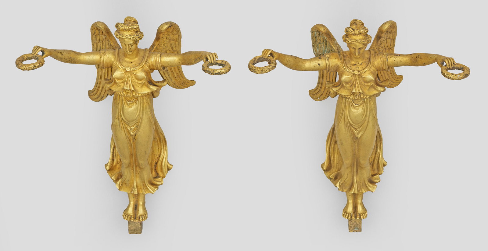 Paar figurale Empire-Möbeldekorationen: Bronze, fein ziseliert und feuervergoldet. Vollplastisch gestaltete Darstellung von geflügelten Genien in antikisierendem Gewand, zwei Lorbeerkränze emporhaltend. Unterhalb gekanteter Montagesockel.