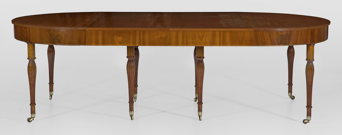 Großer Directoire-Ausziehtisch: Mahagoni, massiv und furniert. Bombierte Zarge auf mehrfach gegliederten, teilw. godronierten Balusterbeinen mit Messingrollen. Umlaufend der Zarge schmale Fadeneinlage. Gering überstehende, profilie