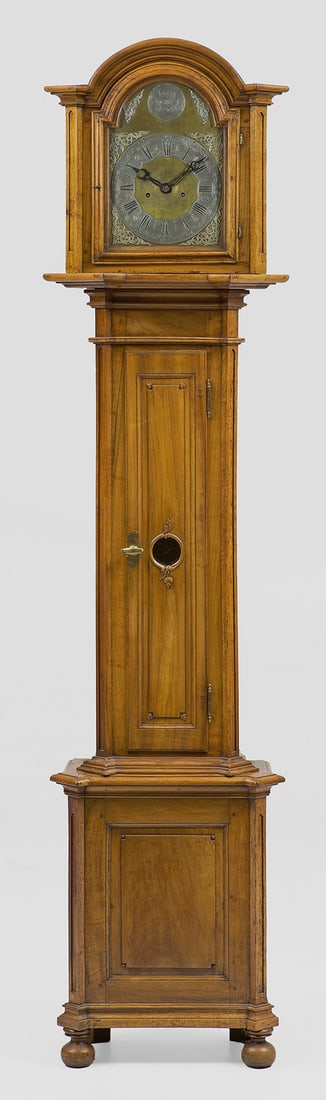 Louis XVI-Bodenstanduhr: Nussbaum, teilw. reliefplastisch geschnitzt. Schlanker, eintüriger Korpus, flankiert von schräggestellten Ecklisenen auf hohem Podestsockel. Eintürig verglaster Uhrenkopf mit segmentförmigem Profi