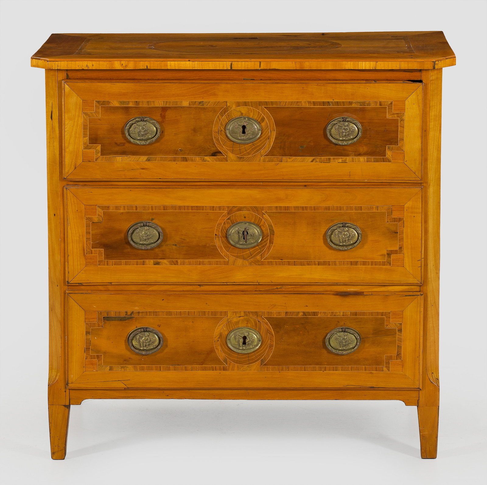 Kleine Louis XVI-Kommode: Kirschbaum, furniert. Gerader, rechteckig angelegter Korpus, flankiert von schräggestellten Ecklisenen, auf konischen Vierkantbeinen. In der gegliederten Front drei Schubladen. Gering überstehende D