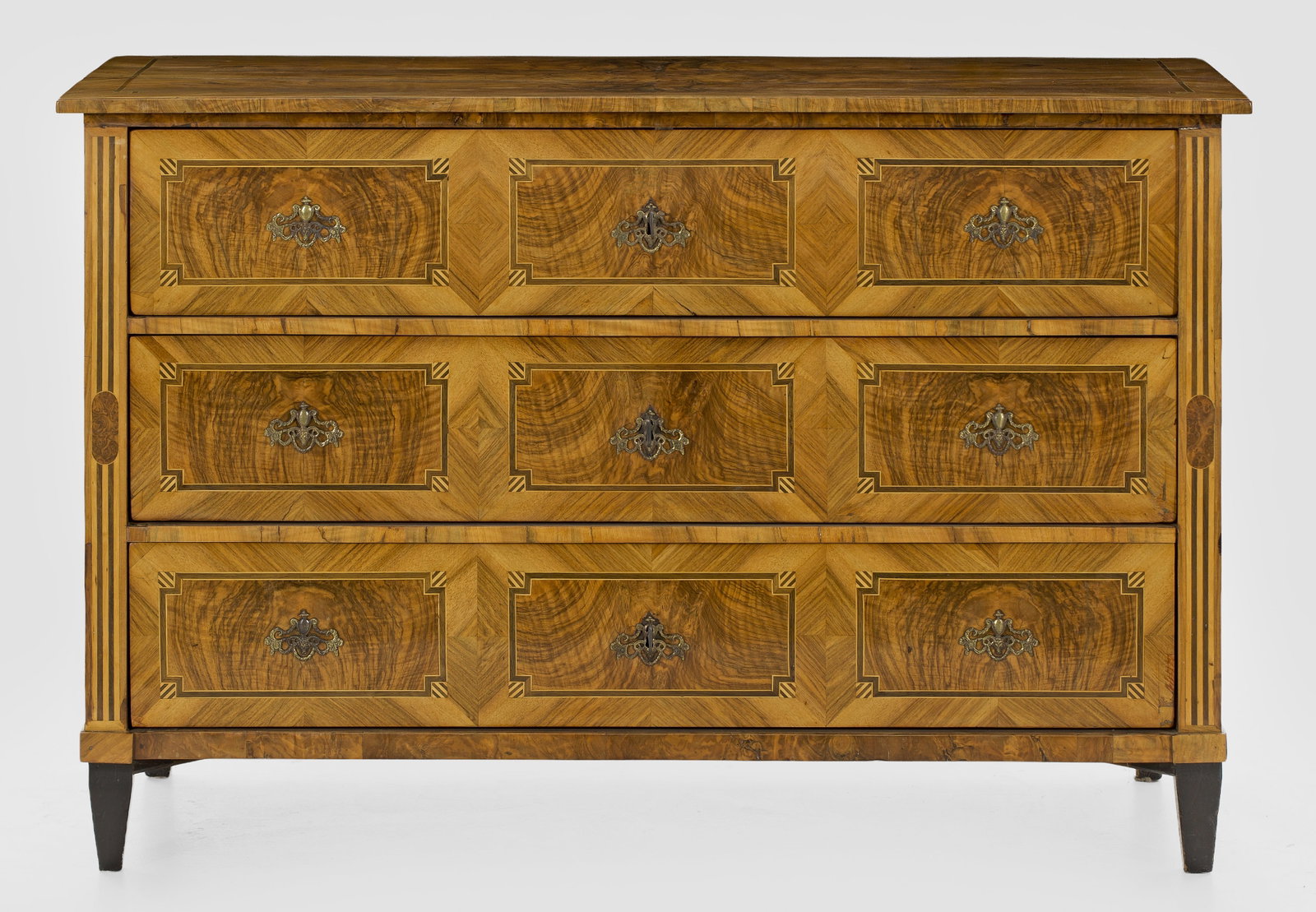 Große Louis XVI-Kommode: Nussbaum, furniert sowie Einlagen aus unterschiedlichen Hölzern. Gerader, querrechteckig angelegter Korpus auf schmaler Zarge und konischen Vierkantbeinen. In der gegliederten Front drei breite Schub