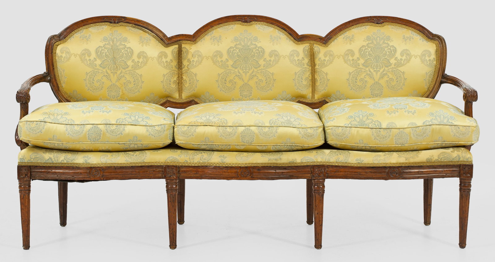 Louis XVI-Bank: Nussbaum, reliefplastisch geschnitzt. Bombierte Zarge auf konischen Balusterbeinen. Geschweifte, blattreliefierte Stütze für gebogte Armlehnen. Leicht gewölbte Rückenlehnenrahmung mit segmentförm