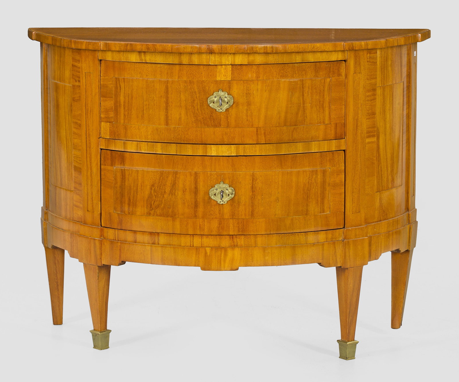 Louis XVI-Demilune-Kommode: Helles Mahagoni, furniert. Bombierter Korpus auf konischen, teilw. verkürzten Vierkantbeinen. In der gegliederten Front zwei Schubladen, flankiert von flachen Pilastern. Dem Korpus entsprechende, ger