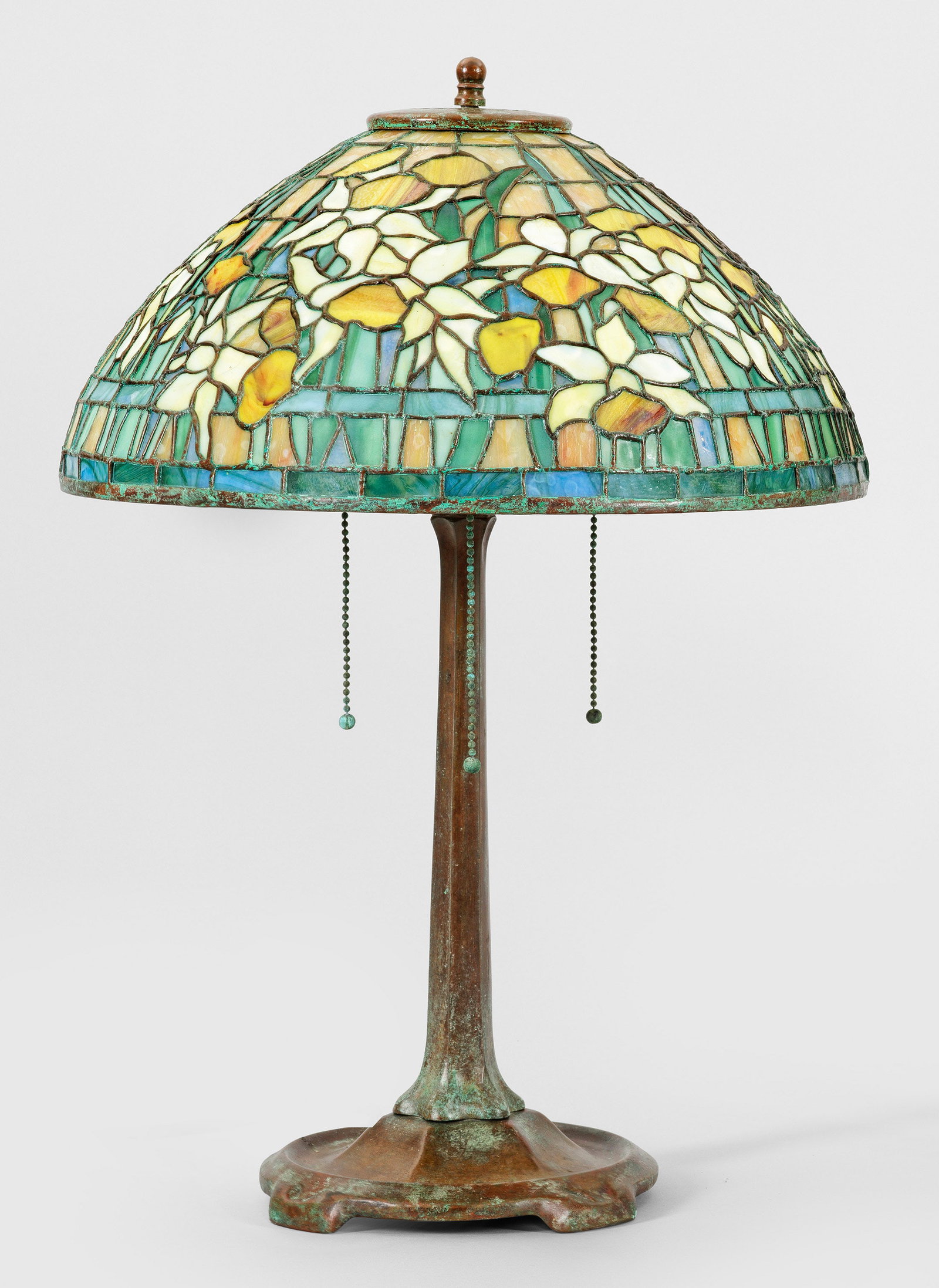 Tischlampe im Tiffany-Stil: 3-flg.; Bronze, grün-braun patiniert sowie farbiges Glas in unterschiedlichen Gelb-, Grün- und Blautönen. Über geschweift ansteigender Basis sich konisch verjüngender Schaft als Träger für Leuc