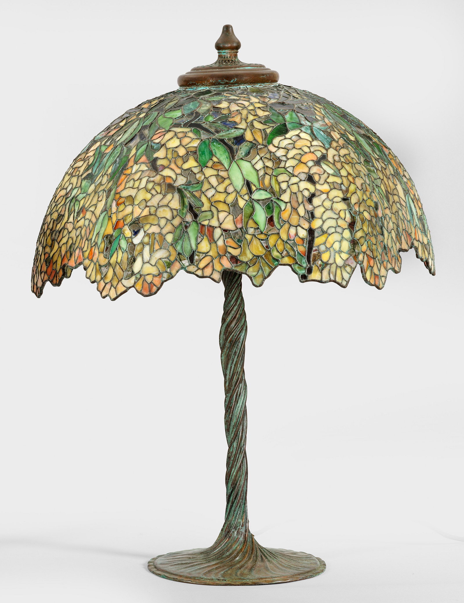 Große Tischlampe im Tiffany-Stil: 4-flg.; Bronze, grün-braun patiniert sowie farbiges Glas in unterschiedlichen Gelb- und Grüntönen. Flache, geschweift ansteigende Basis in gestreckten, in sich gedrehten Schaft als Träger für Leu