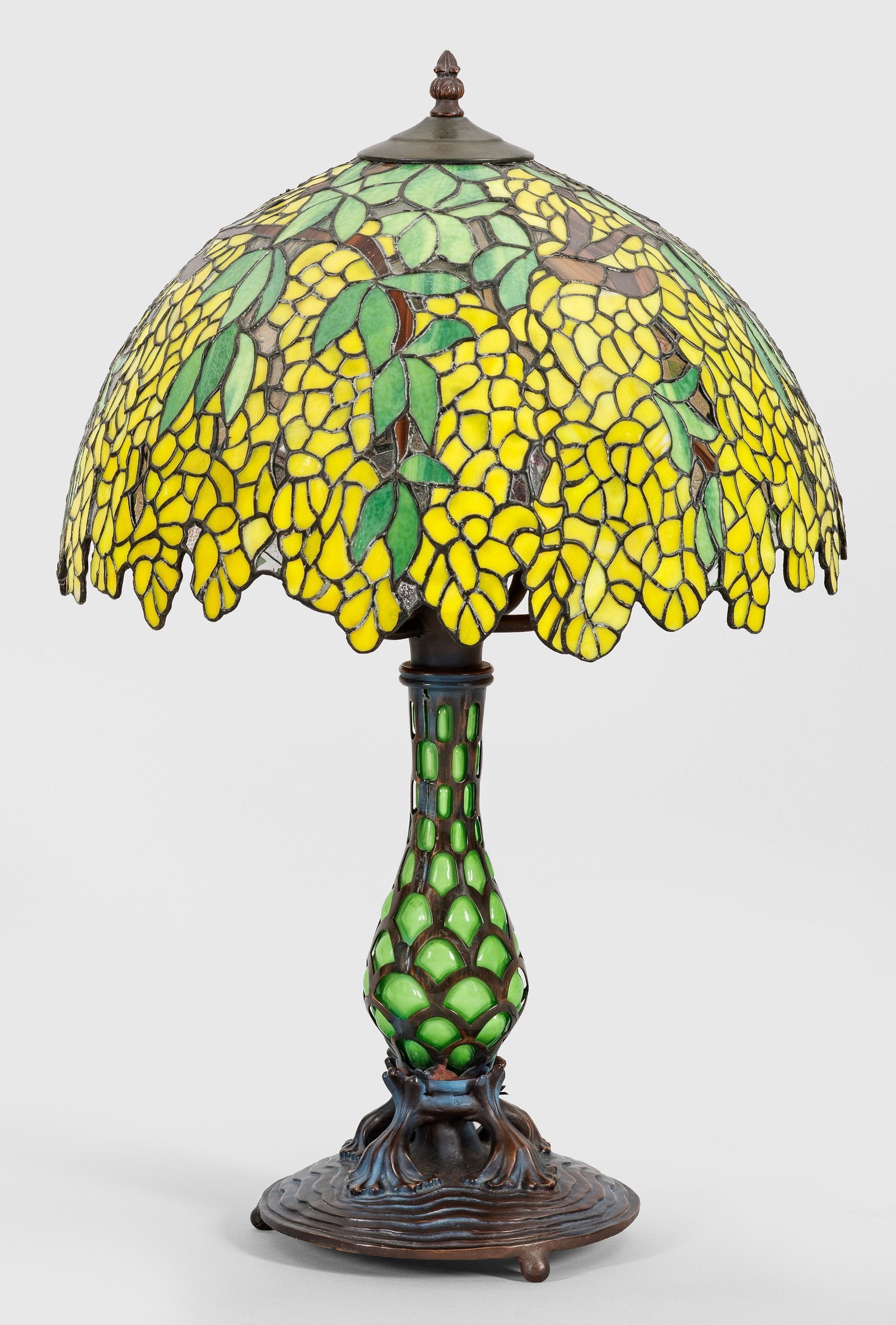 Tischlampe im Tiffany-Stil: 6-flg.; Bronze, dunkel patiniert sowie farbiges Glas in Gelb- und Grüntönen. Über geschweift ansteigendem, profilierten Sockel plastische Blattelemente sowie balusterförmiger, durchbrochen gearbei