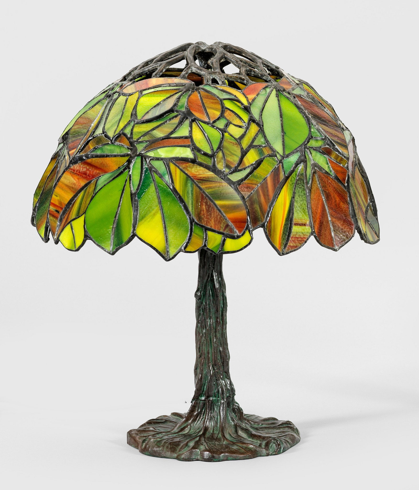 Tischlampe im Tiffany-Stil: 3-flg.; Bronze, patiniert sowie farbiges Glas in unterschiedlichen, leuchtenden Grüntönen. Naturalistisch gestalteter Schaft in Form eines Baumstamms als Träger für Leuchtmittel. Kuppelförmiger L