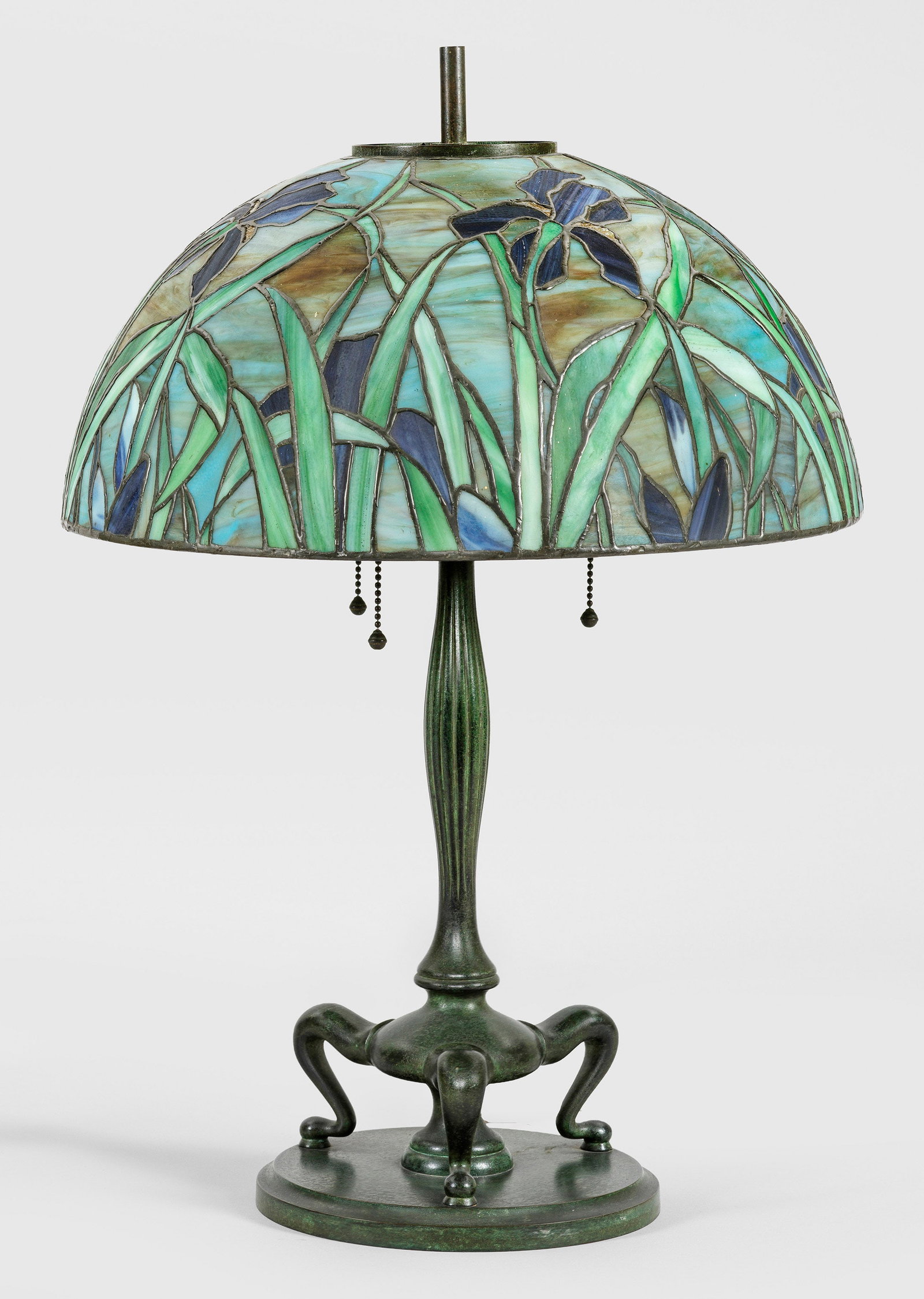 Tischlampe im Tiffany-Stil: 3-flg.; Bronze, grün patiniert sowie farbiges Glas in unterschiedlichen Grün- und Blautönen. Über flachem Rundsockel geschwungene Volutenmotive und godronierter Balusterschaft als Träger für Leu