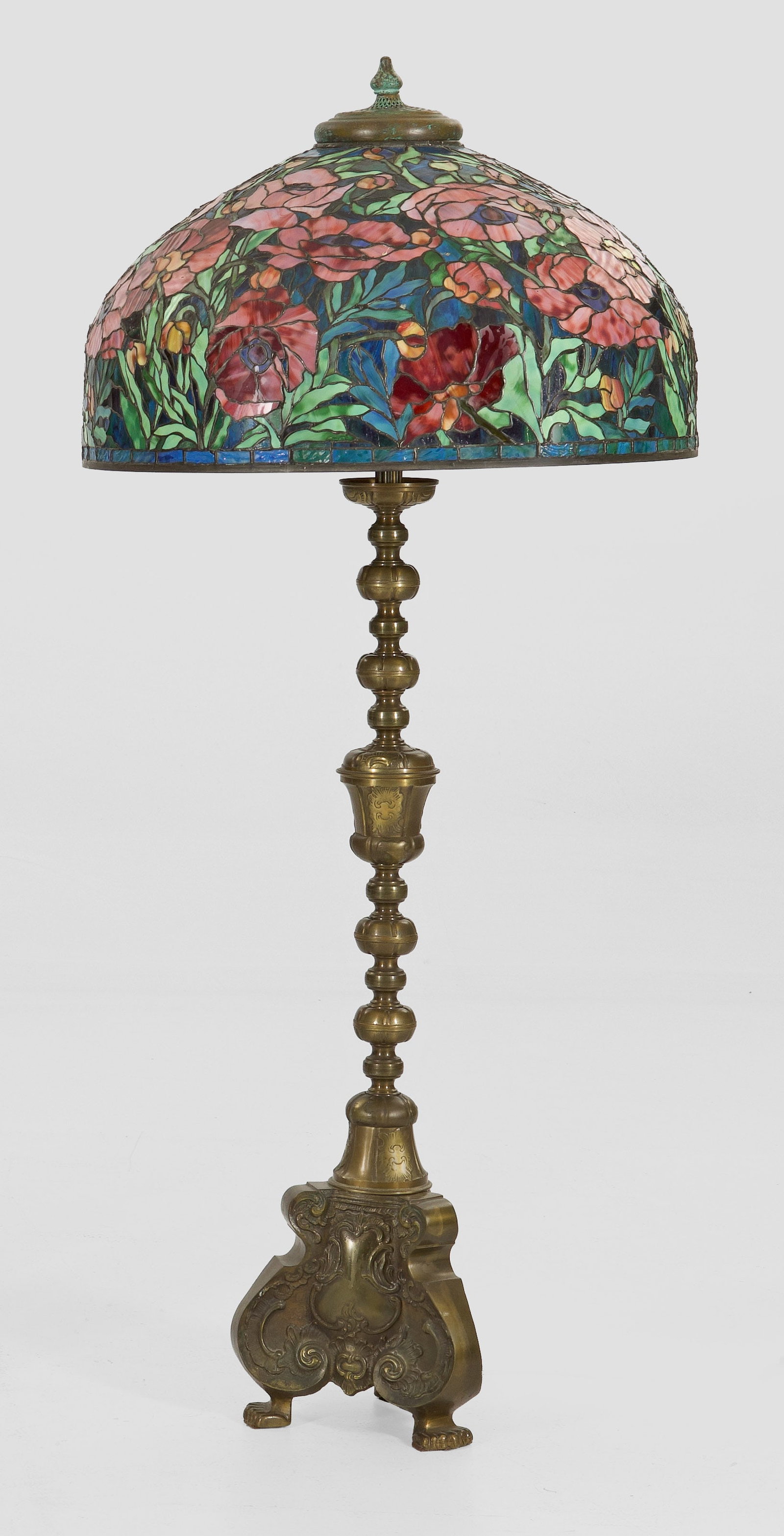 Bodenlampe mit Lampenschirm im Tiffany-Stil: 6-flg.; Messing sowie farbiges Glas in unterschiedlichen blau-, Grün- und Rosétönen. Über mehrfach gegliedertem, reliefiertem Balusterschaft auf gedrungenen Volutenmotiven kuppelförmiger