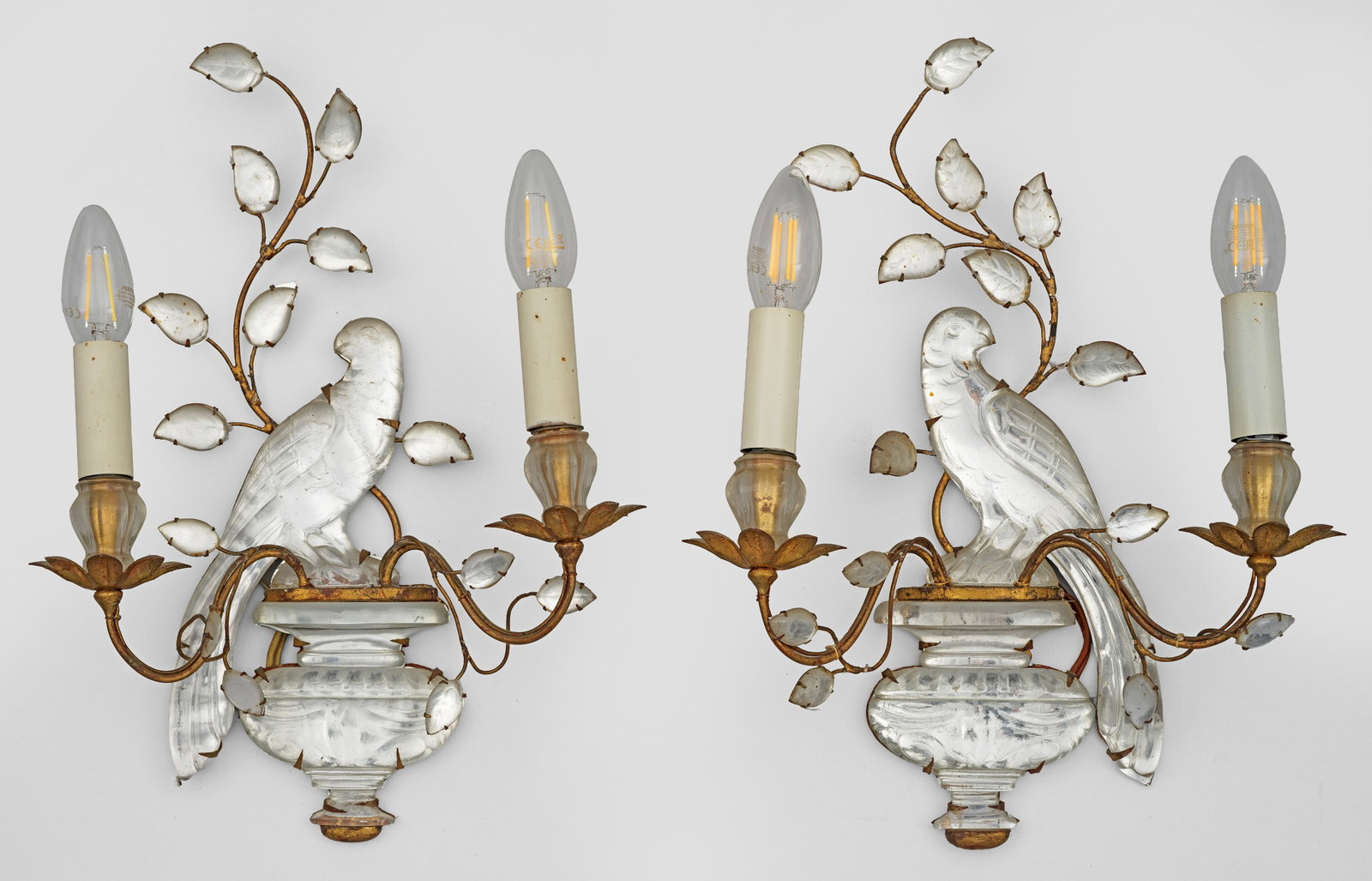 Paar Wandlampen von Maison Baguès: Gegenstücke. 2-flg.; Metall, vergoldet sowie farbloses, mit Silberfolie hinterlegtes Glas. Balusterförmiges Wandschild mit erhaben reliefiertem Papagei und geschwungenem Zweig. Davon ausgehend korre