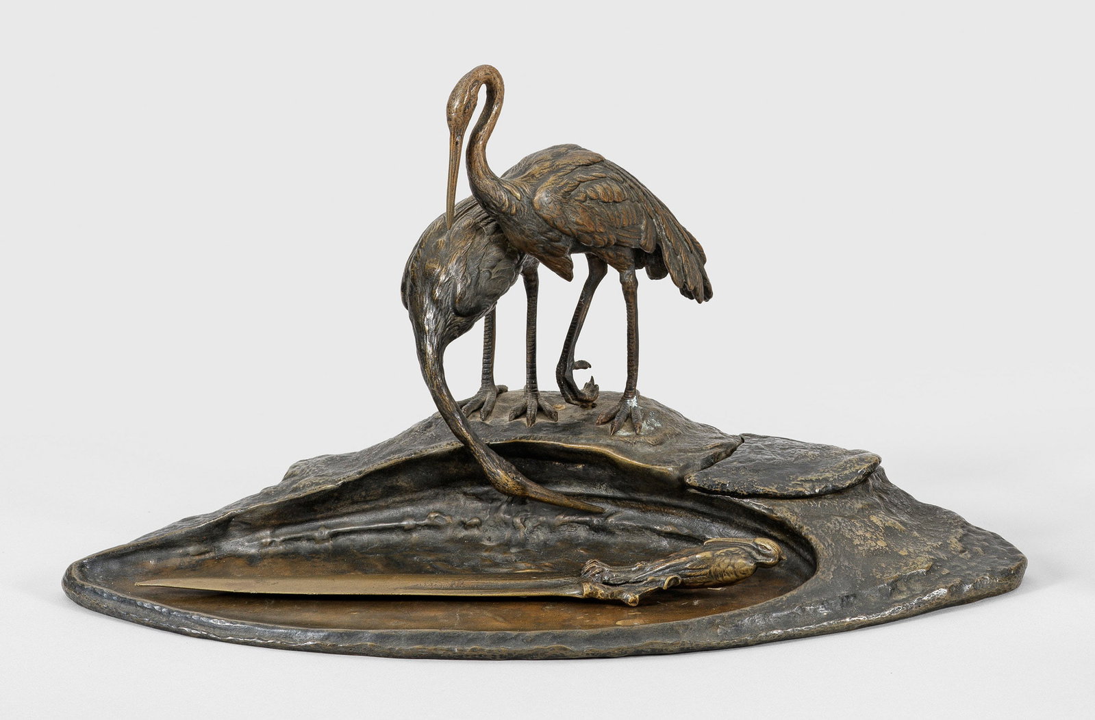 Skulpturales Jugendstil-Schreibset von Ruffino Besserdich: 2-tlg.; Bronze, dunkel patiniert. Sichelform. Am Ufer stehend zwei trinkende Reiher mit fein reliefiertem Gefieder. Dazu Brieföffner mit Griff in Gestalt des Vogels. Seitlich verdeckter Glaseinsatz. 