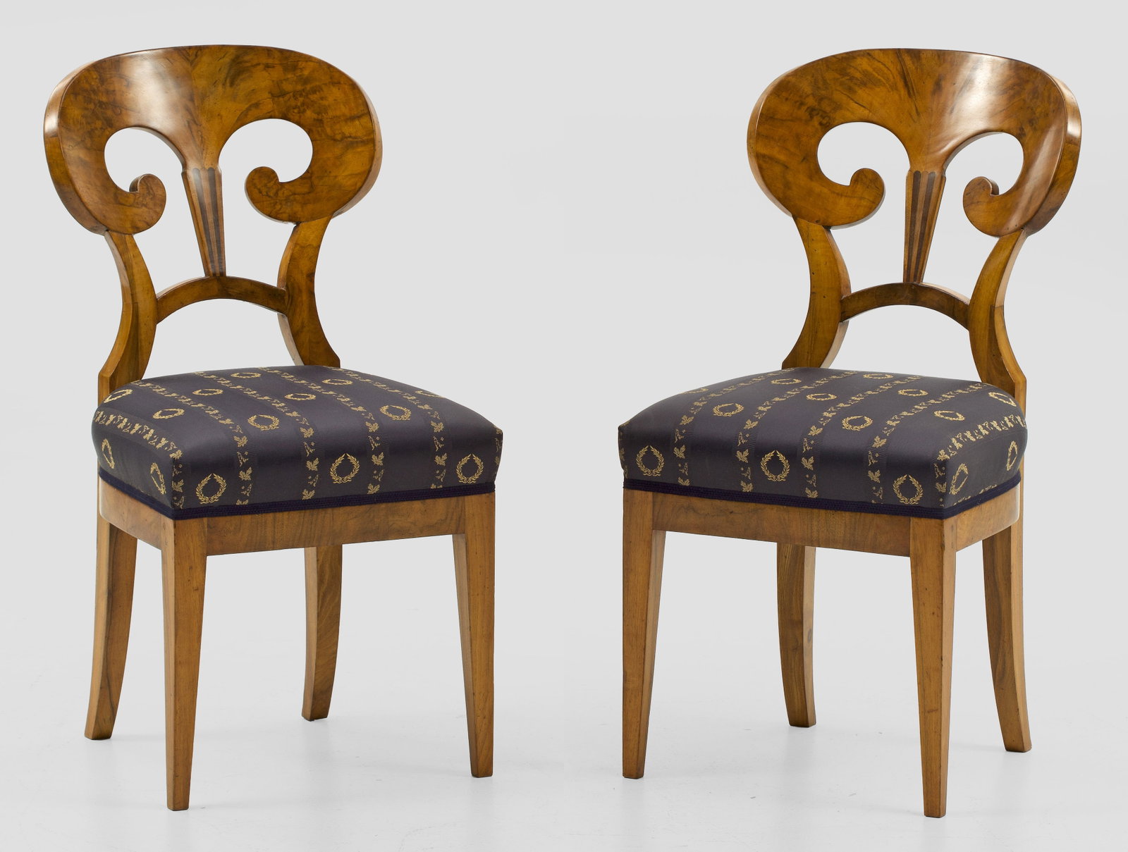 Paar Biedermeier-Stühle: Nussbaum, furniert. Bombierte Zarge auf konischen Vierkantbeinen. Gewölbte, schaufelartige Rückenlehne aus stilisierten Voluten. Sitzfläche gepolstert und bezogen. Sitz-H. 45 cm. A pair of South Ge