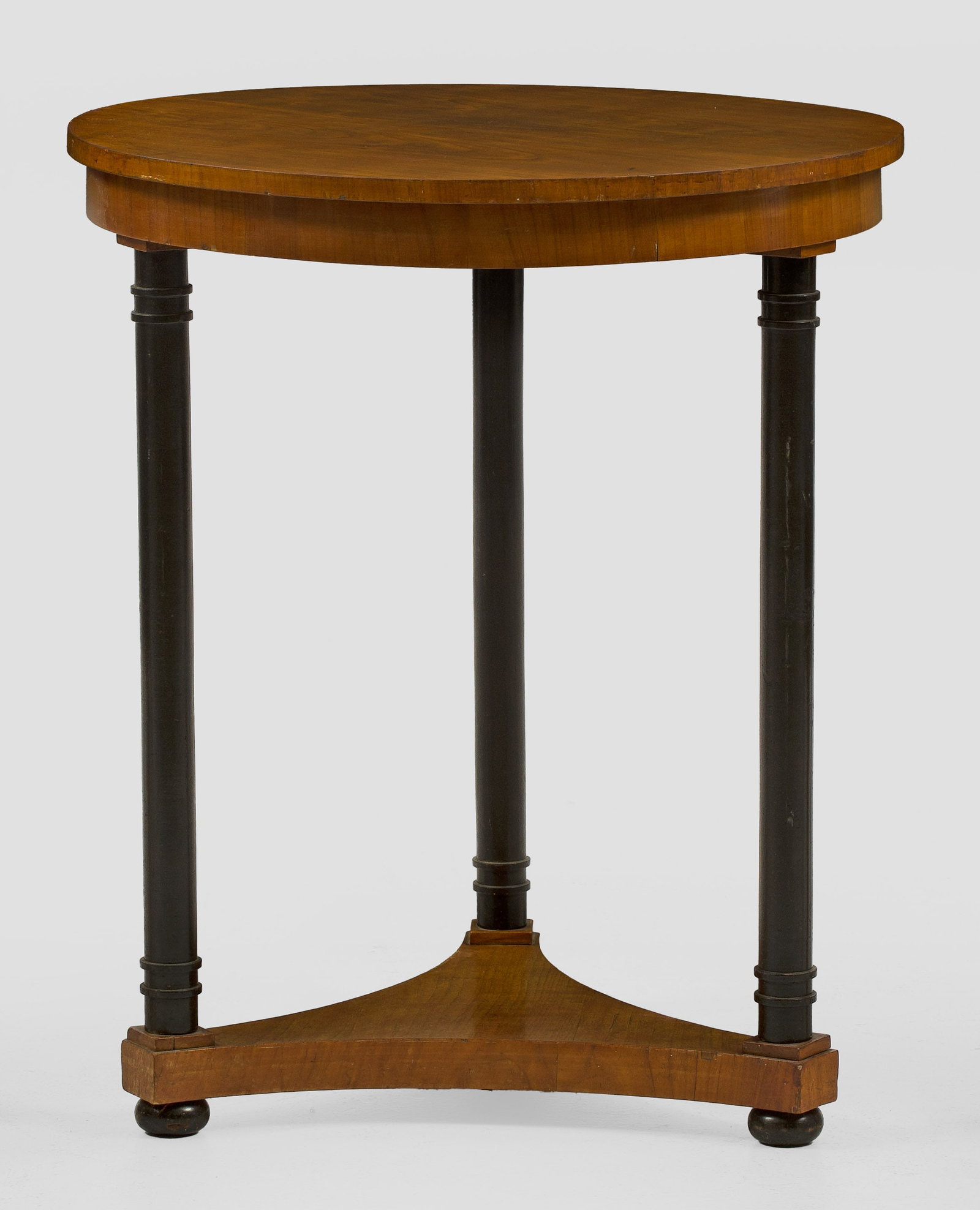 Biedermeier-Beistelltisch: Kirschbaum, furniert und teilw. ebonisiert. Über dreiseitig konkav eingezogener Sockelplatte zylinderförmige Schäfte als Träger für bombierte Zarge und runde Deckplatte. H. 76 cm. D. 63,5 cm. A S
