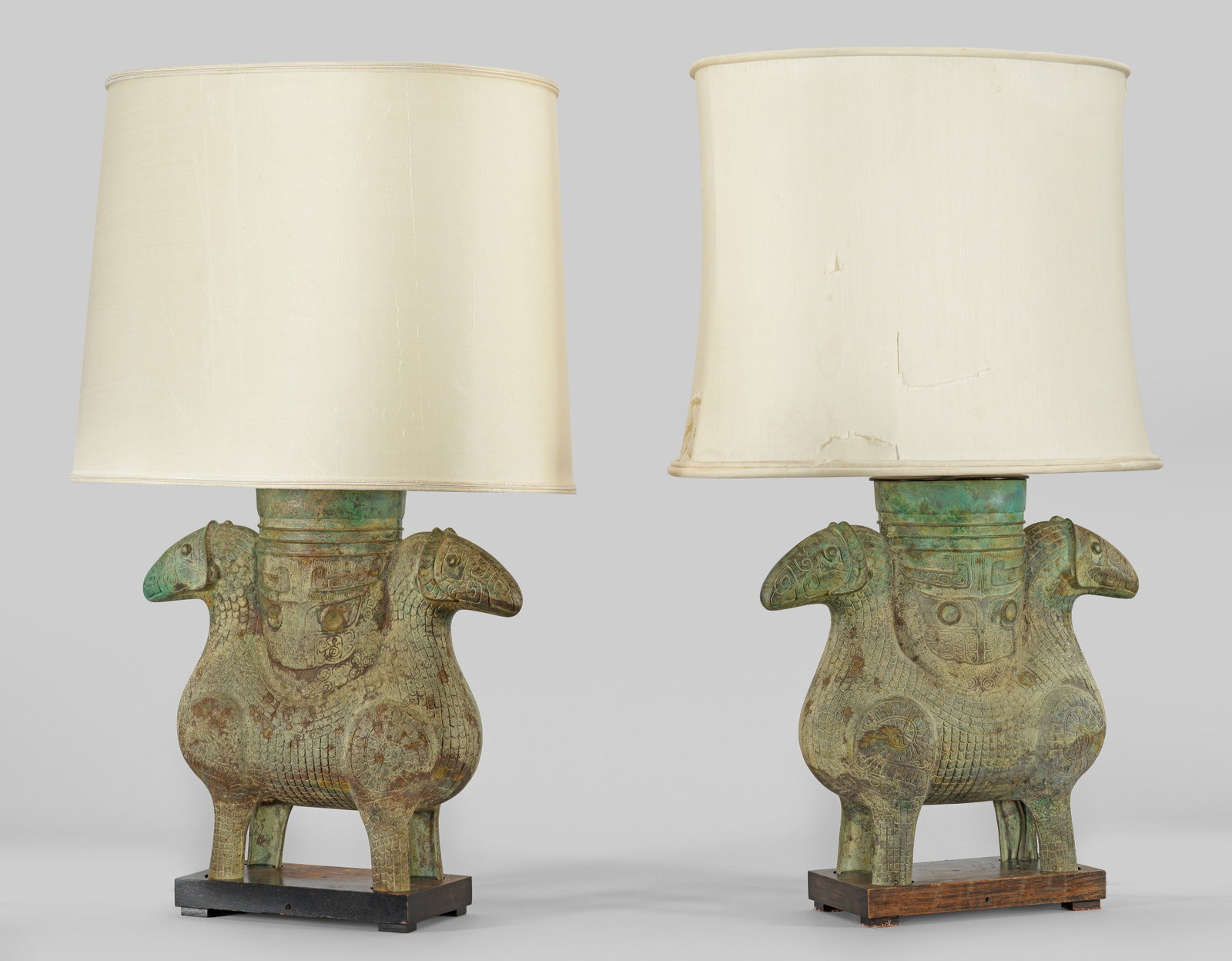 Paar Tischlampen im Stil der Shang Dynastie: 2-flg.; Bronze, grün patiniert sowie Holz und Metall. Über flachem, rechteckigen Sockel vollplastische Darstellung eines sog. Doppelwidders nach einem historischen Vorbild aus der Shang Dynasty. In 