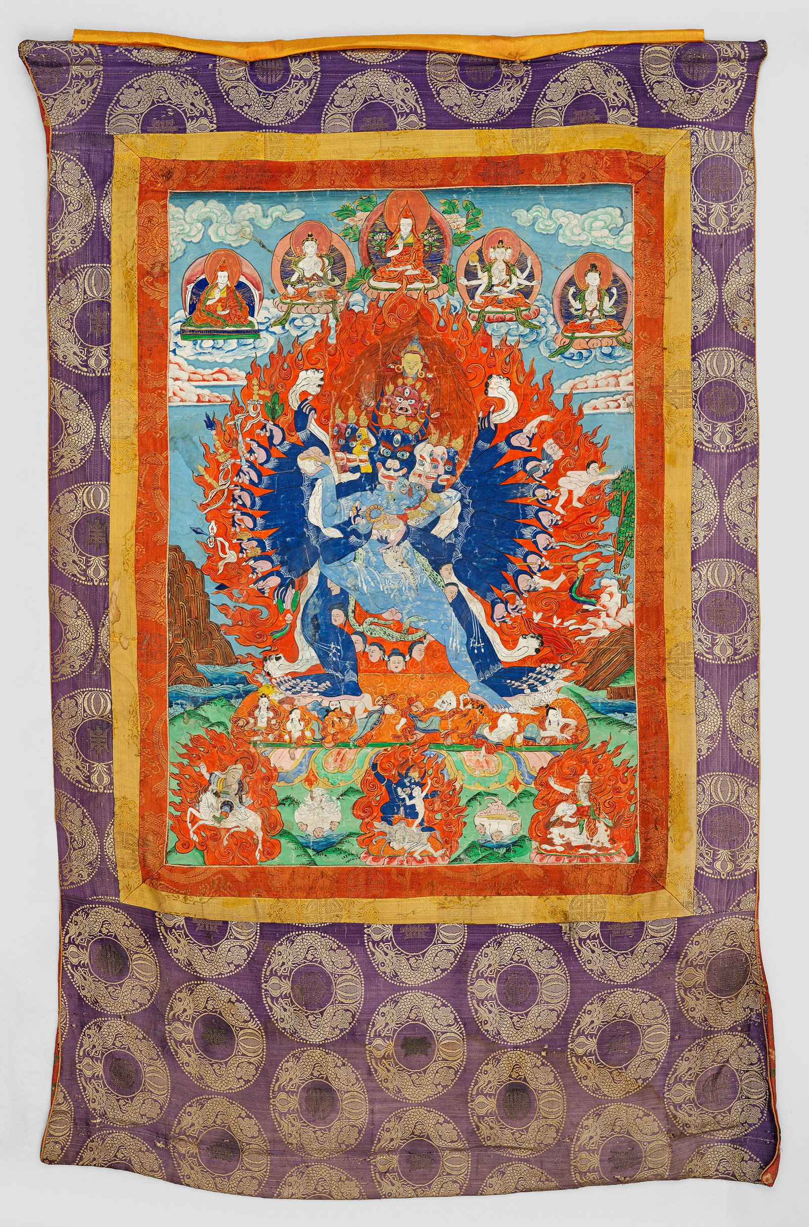 Tibetische Thangka: Gouache auf Leinwand. Rechteckige Form mit dreifach eingefasstem, zentralen Bildfeld. Darin in weitläufiger Küstenlandschaft szenische Darstellung der von Flammen umzogenen Gottheit Yamantaka