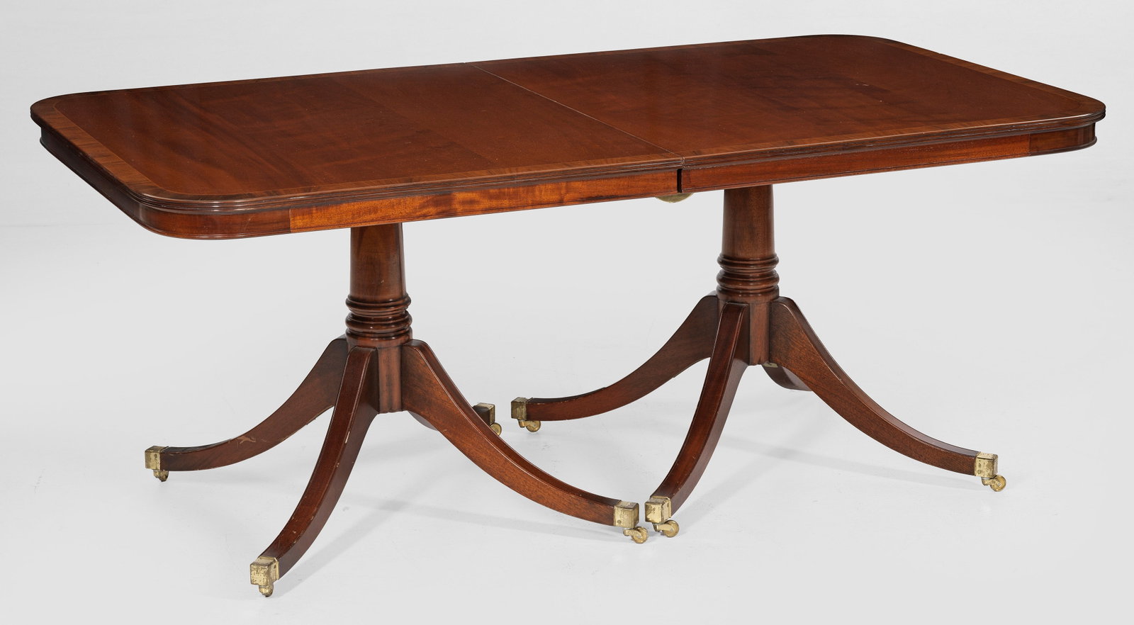 Ausziehtisch im Regency-Stil: Mahagoni, furniert. Konische Balusterschäfte aus geschweiften Beinen mit Messingrollen als Träger für gerade Zarge und gering überstehende, profilierte Deckplatte mit gerundeten Ecken. Dazu zwei E
