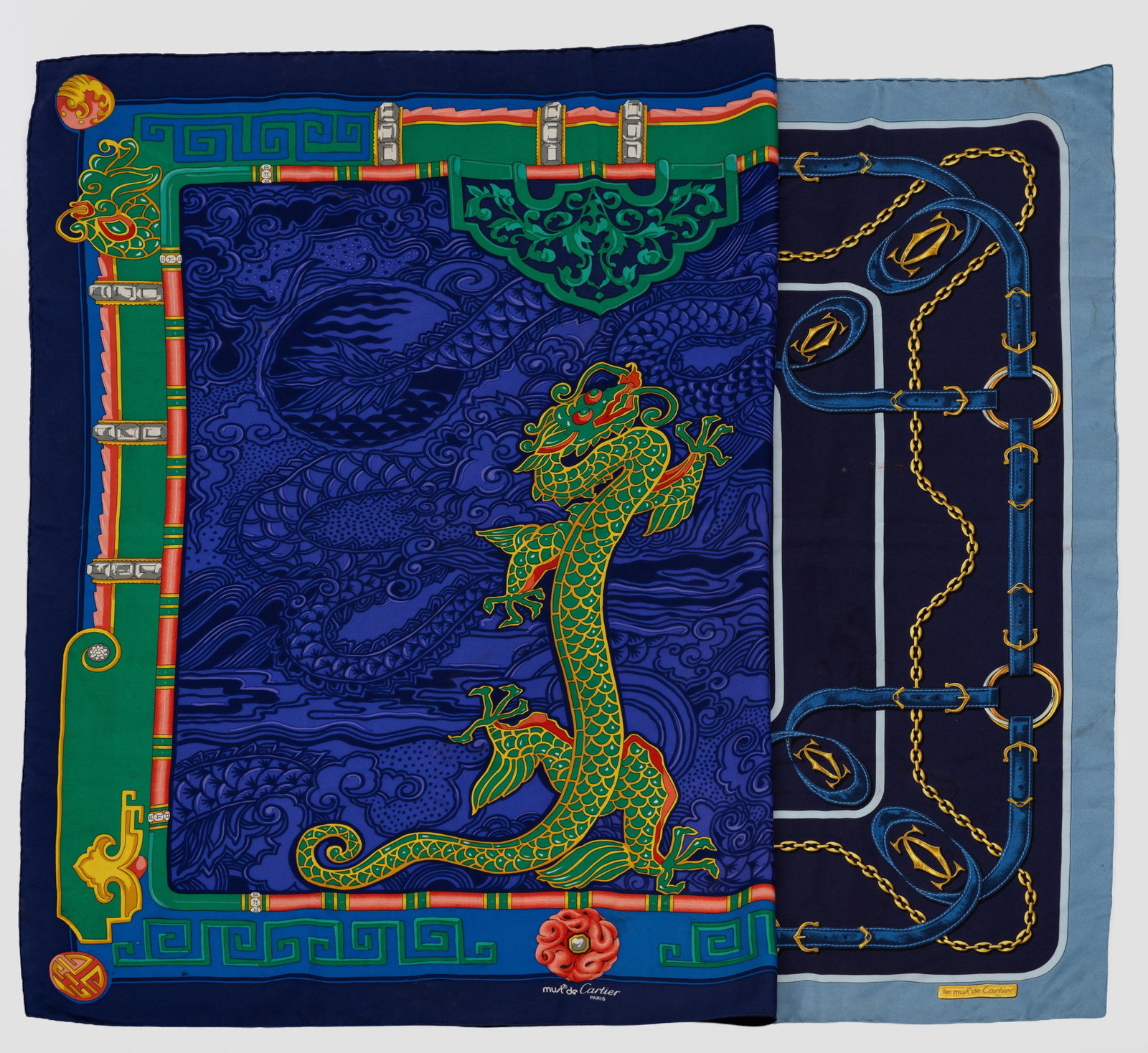 Zwei "Must de Cartier"-Seidentücher von CARTIER: Seide. Quadratisches Tuch. Auf blauem Fond Dekor aus Zügeln, Gebissringen und Schnallen bzw. aus asiatischen Wolken- und Architekturdarstellungen mit zentralem Drachen. "Must de Cartier"-Schriftzug. 