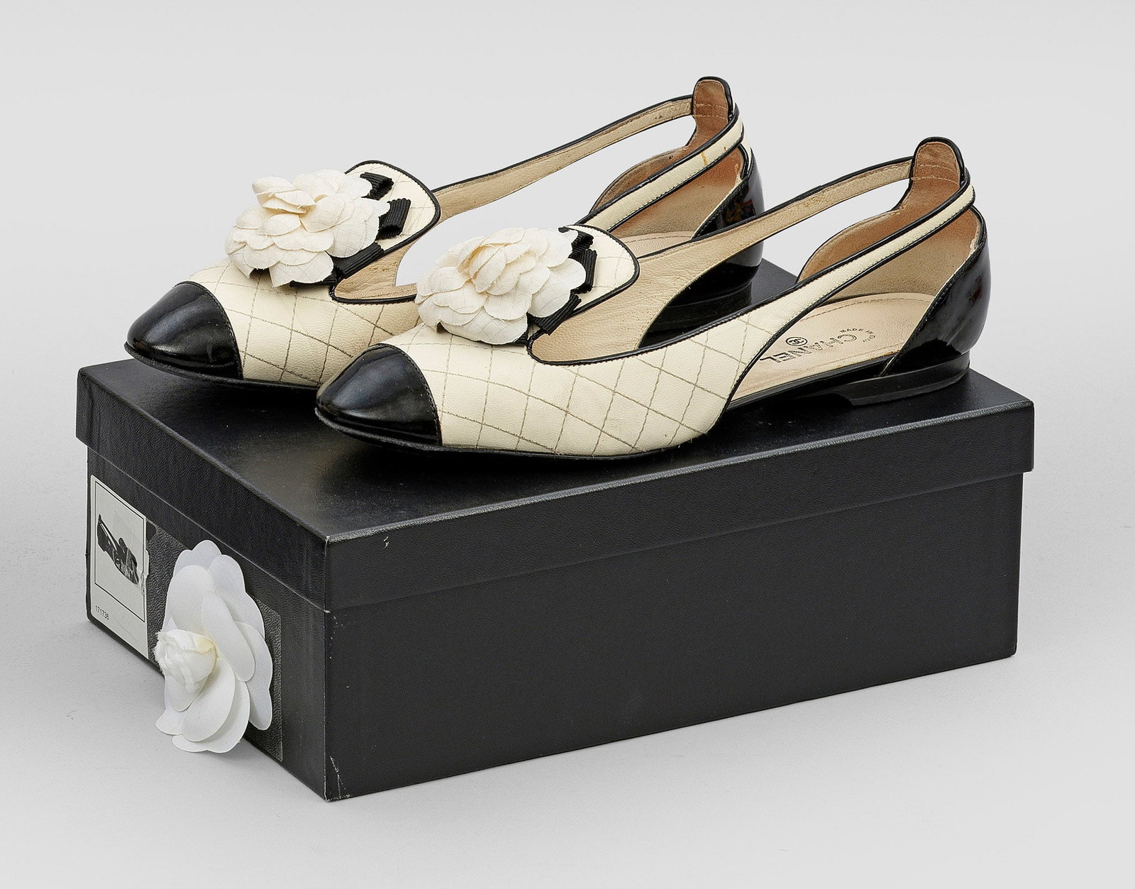 Paar Slingback-Flats von CHANEL: Cremeweißes Leder mit charakteristischer Rautensteppung und schwarzes Lackleder. Klassisch geschnittener Damenschuh mit farblich abgesetzter Spitze und Dekor aus aufgelegter, textiler Kamelienblüte.