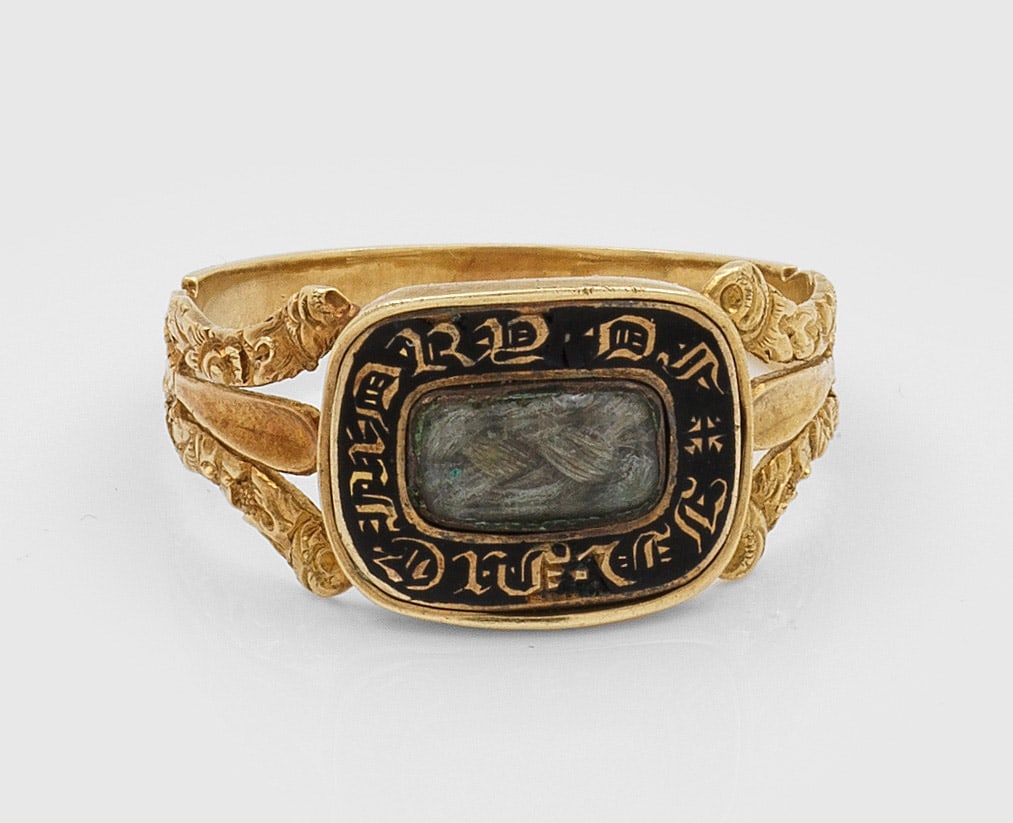Viktorianischer Memorial-Ring von 1843: Gelbgold, 18 ct., teilw. schwarz emailliert. Floral reliefierte Ringschiene, zentrales verglastes Medaillon mit Inschrift und gelochtem Haareinsatz, verso gravierte Inschrift von 1843. Gew. ca. 3,30 g