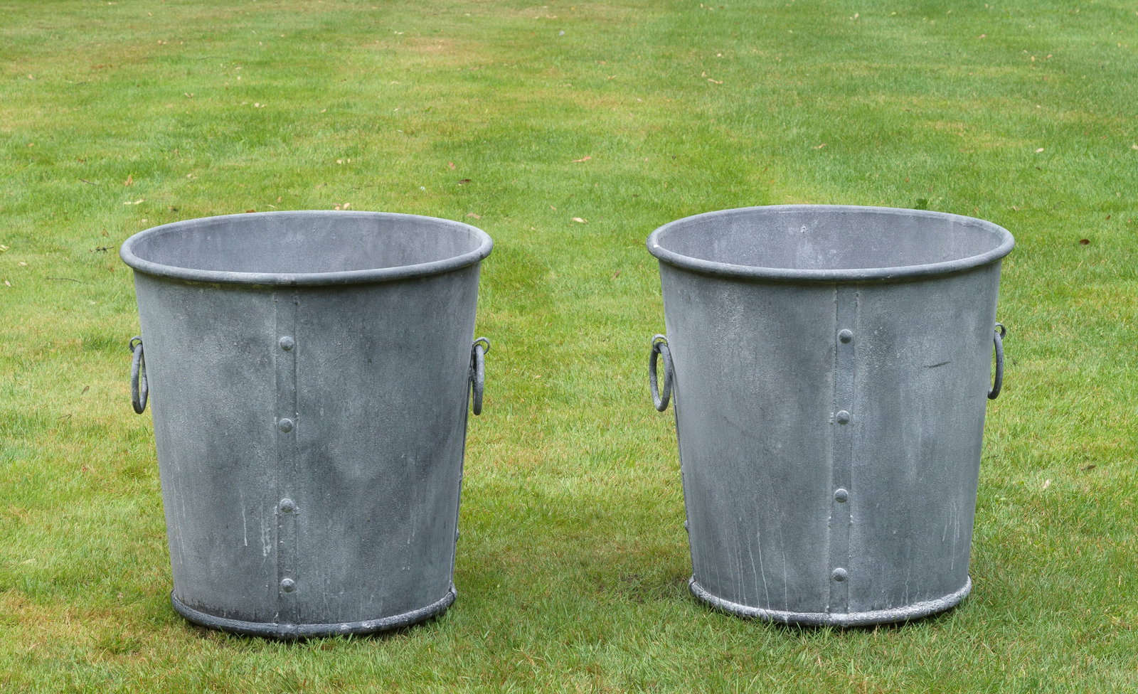 Paar große Palmenkübel: Metall, grau patiniert. Zylinderförmig sich erweiternder, glatter Korpus mit Wulstrand. Seitliche Nutleisten mit hängenden Ringen. H. 64 cm. A pair of French grey patinated palm tree cachepots. Fran