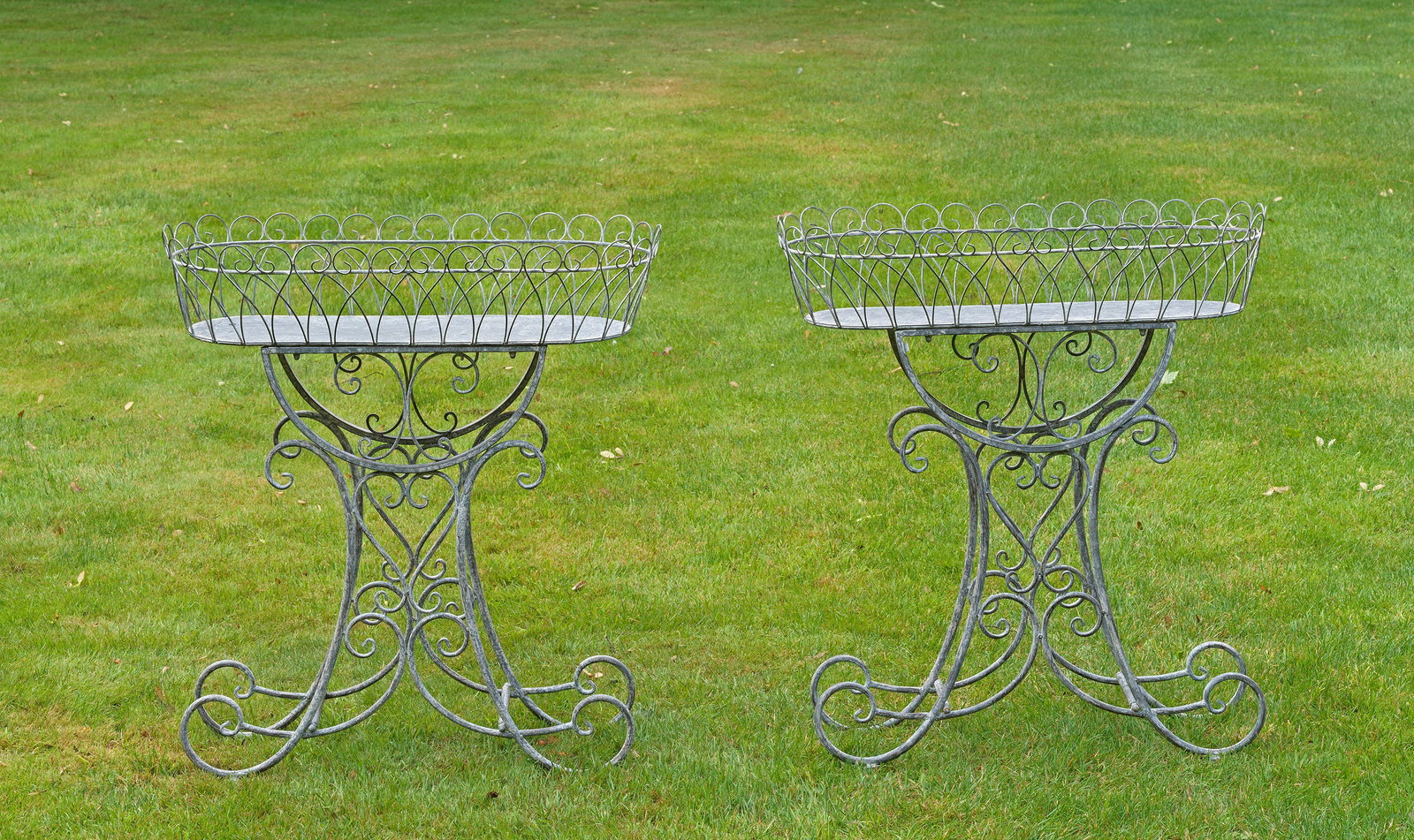Paar Belle Epoque-Blumenbänke: Metall, hellgrau lackiert. Offen gestaltetes Gestell aus gedrehten, volutenförmig eingerollten und ornamental angelegten Rundstäben als Träger für korbähnliche Blumenjardiniere. H. 83 cm. B. 74 c