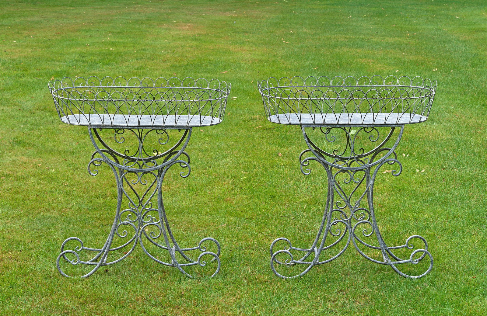 Paar Belle Epoque-Blumenbänke: Metall, hellgrau lackiert. Offen gestaltetes Gestell aus gedrehten, volutenförmig eingerollten und ornamental angelegten Rundstäben als Träger für korbähnliche Blumenjardiniere. H. 83 cm. B. 74 c