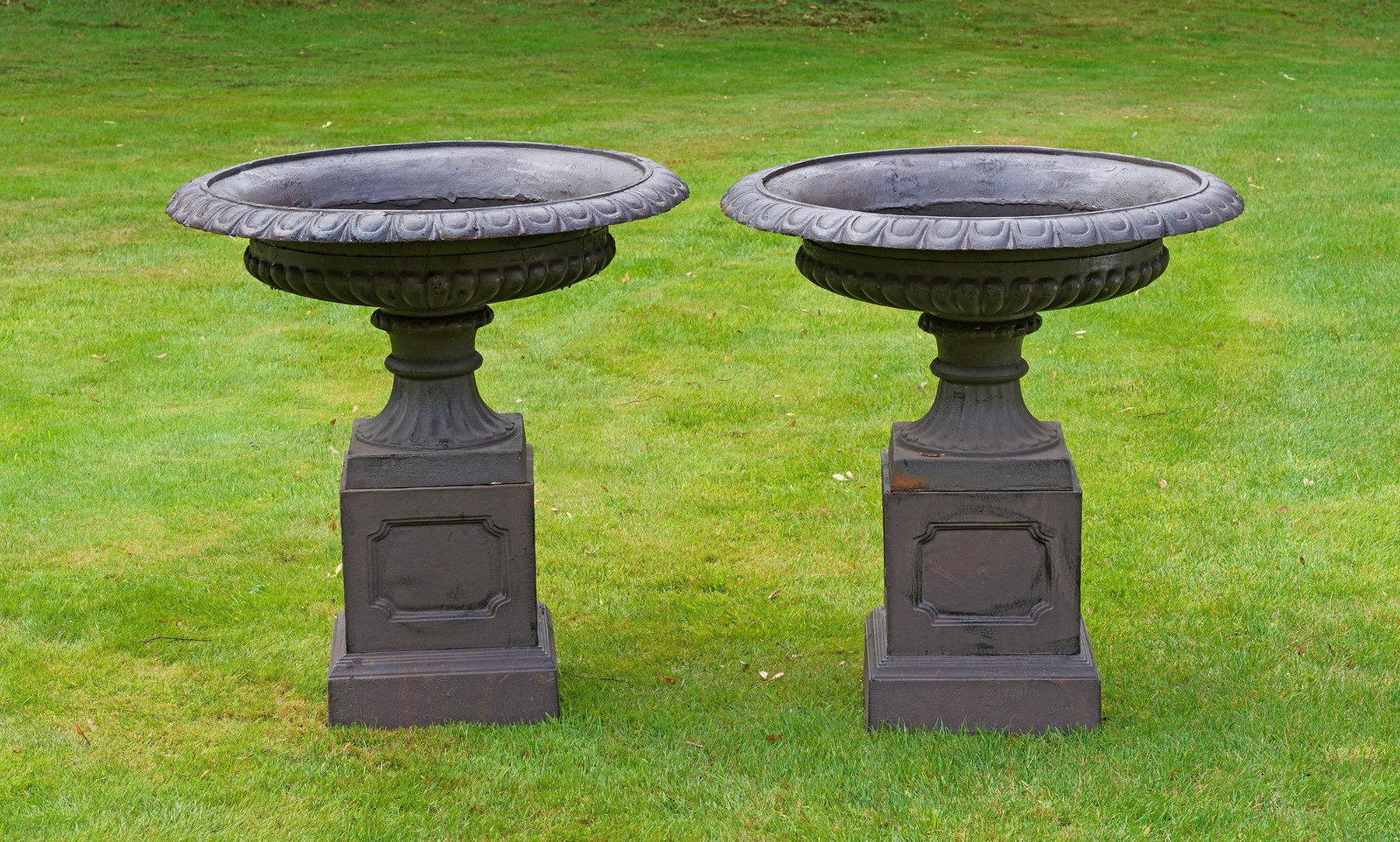 Paar große Parkvasen auf Postamentsockel: Gusseisen, dunkelbraun, in Rostoptik lackiert. Über quadratischer Plinthe flach gewölbter, umlaufend godronierter Korpus mit breitem, weit vorkragendem Lippenrand aus Eierstabfries. Unterhalb gekant