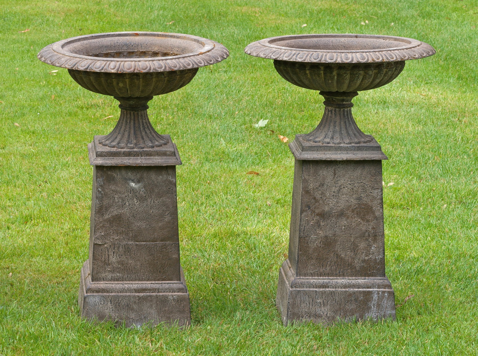 Paar klassische Parkvasen auf Postamentsockel: Gusseisen, dunkelbraun, in Rostoptik lackiert. Über quadratischer Plinthe flach gewölbter, umlaufend godronierter Korpus mit breitem, weit vorkragendem Lippenrand aus Eierstabfries. Unterhalb gestuf