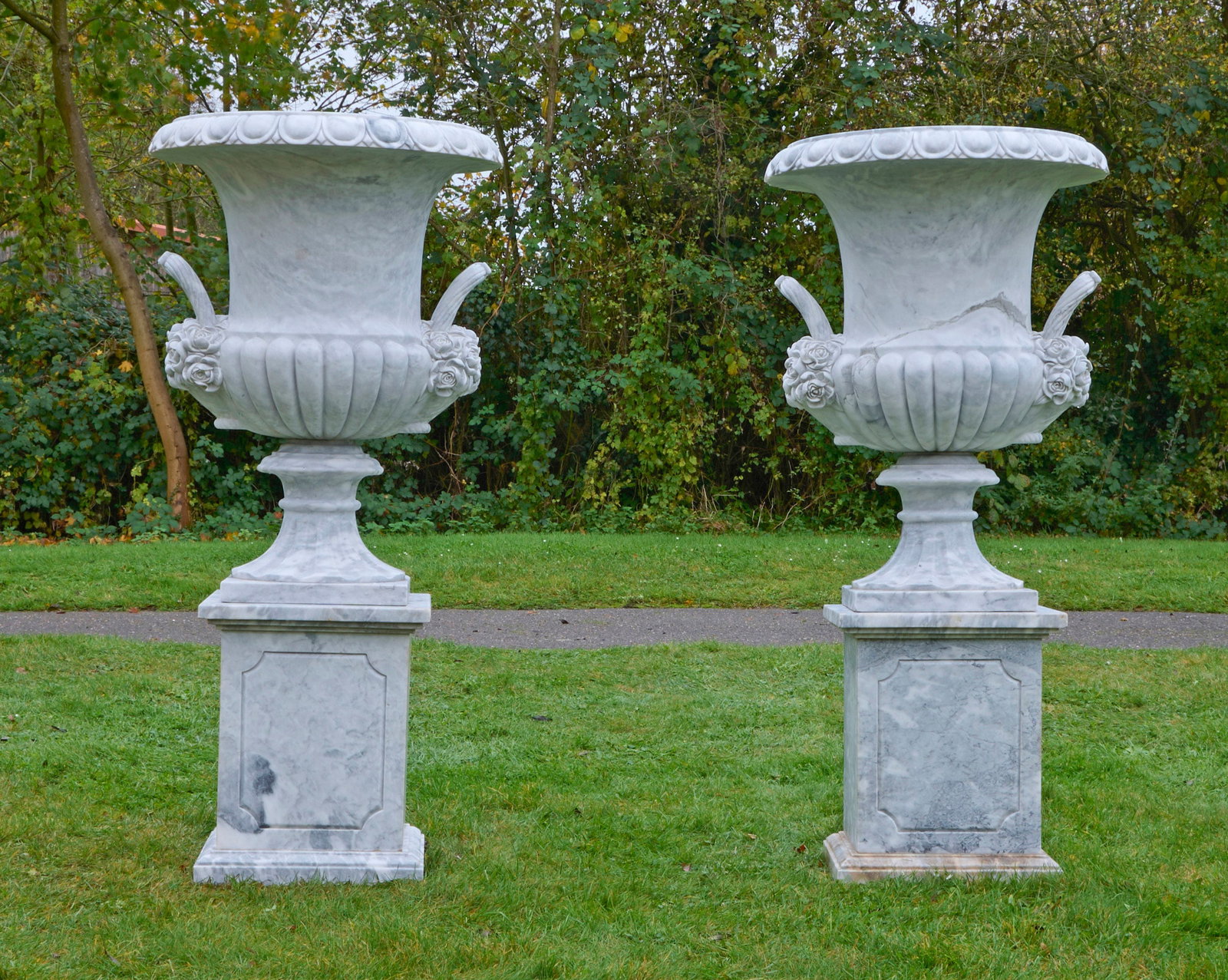 Paar monumentale Parkvasen auf Postamenten: Weißer, hellgrau gemaserter Marmor. Balusterförmige Kratervasen mit teilw. kräftig godronierter Wandung sowie weit ausschwingendem Lippenrand aus breitem Eierstabfries. Seitlich vollplastisch ausge