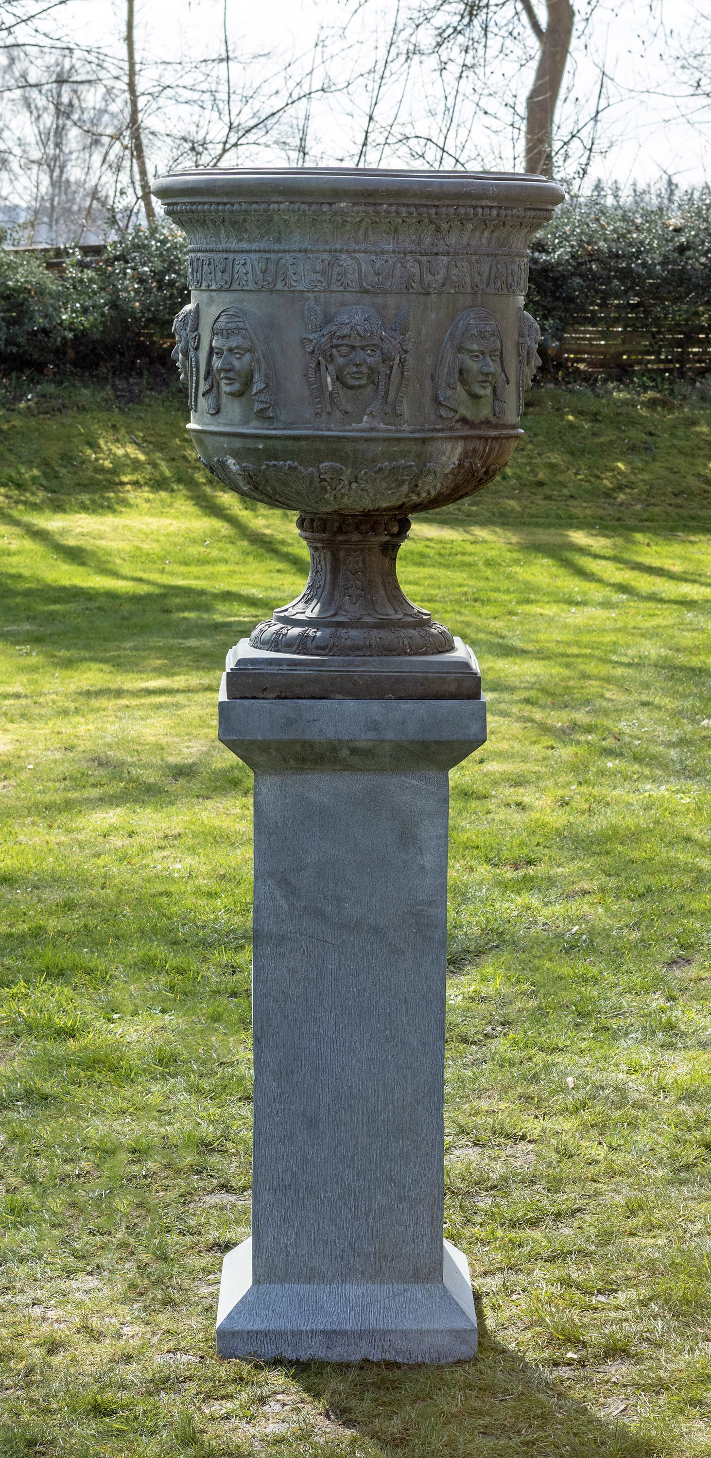 Große Gartenvase auf Podestsockel: Bronze patiniert und Metall. Über flacher, quadratischer Plinthe kelchförmiger, blattreliefierter Korpus mit profiliertem Lippenrand. Umlaufend der Wandung unterschiedliche, erhaben reliefierte Mask
